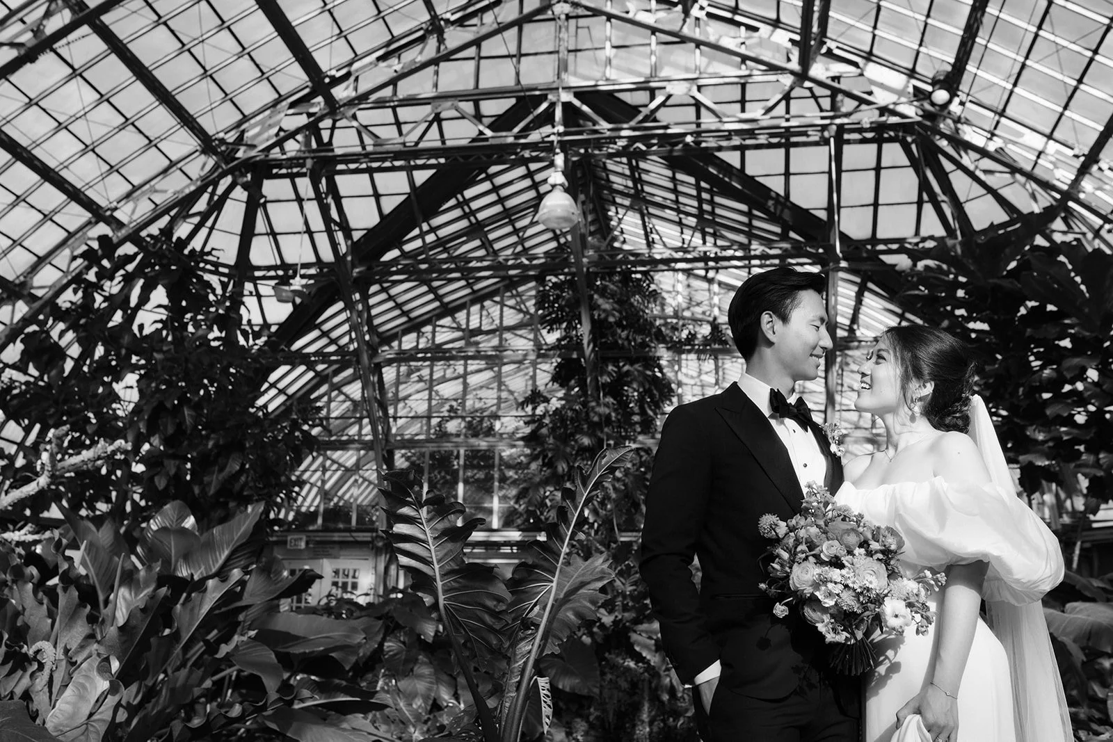 Black and white photo of a smiling bride and groom facing each other with their foreheads nearly touching inside a greenhouse filled with plants and glass ceiling.