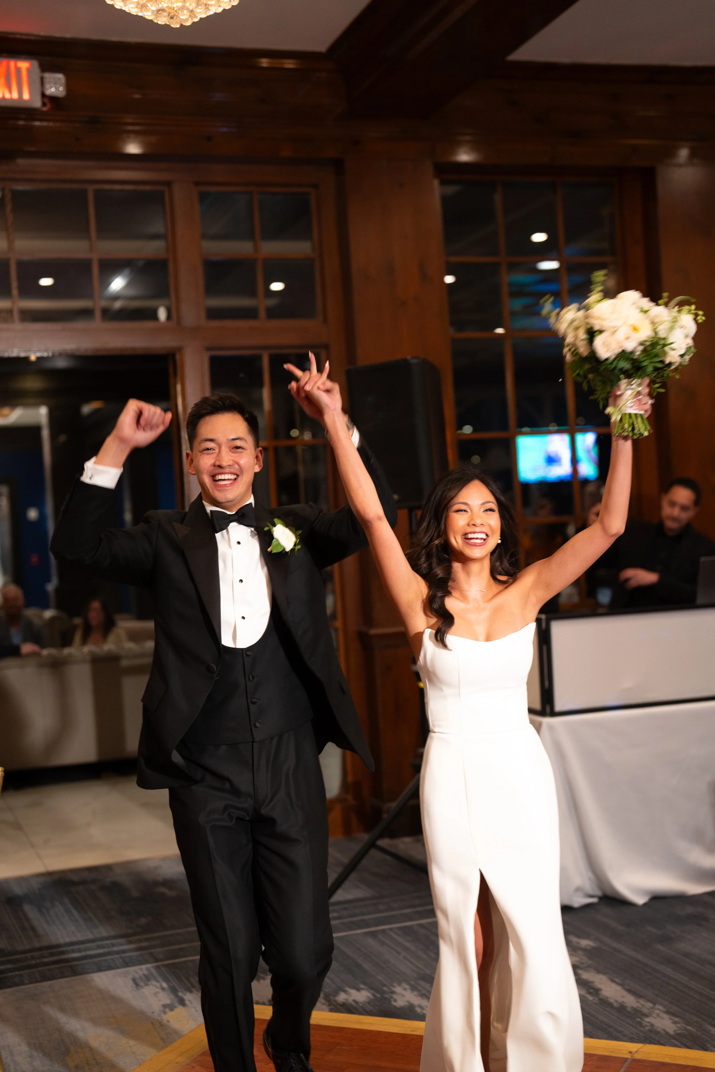 A newlywed couple celebrating at their wedding reception. The groom is dressed in a black tuxedo with a bow tie, joyfully raising his arms, while the bride, wearing a strapless white wedding gown and holding a bouquet of white flowers, is smiling and