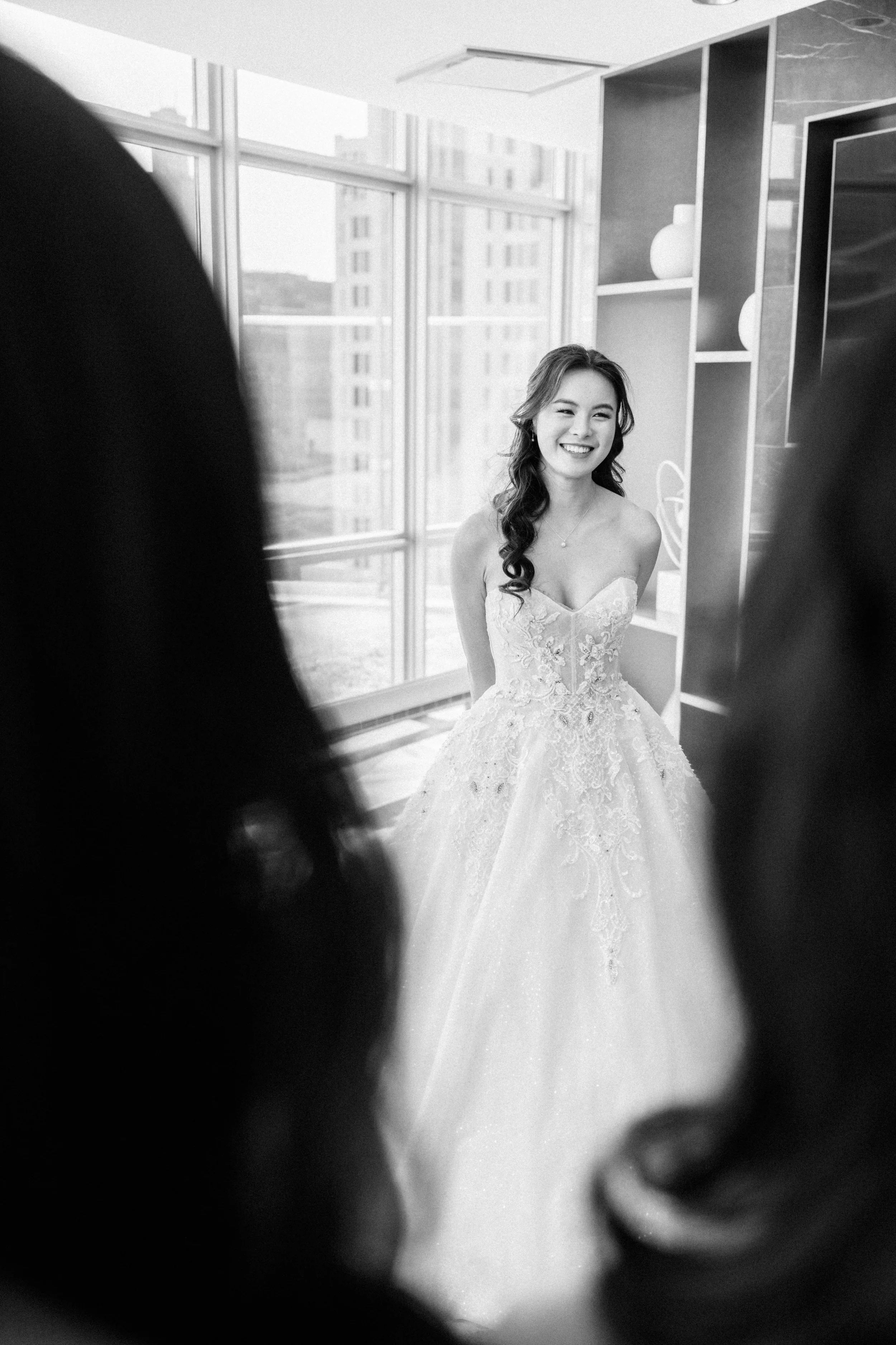 A smiling bride in a wedding dress standing near a window with indoor lighting, with blurred figures in the foreground.