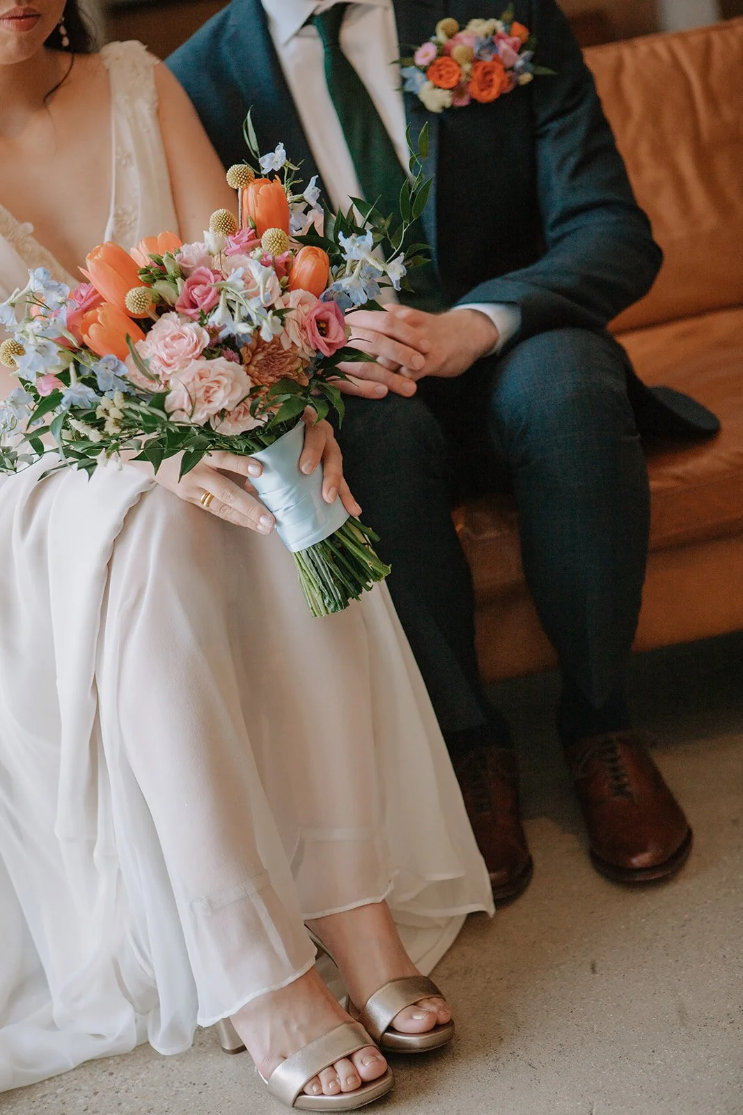 A bride holding a colorful bouquet of flowers, sitting next to a groom in a dark suit with a boutonniere, on a brown couch.
