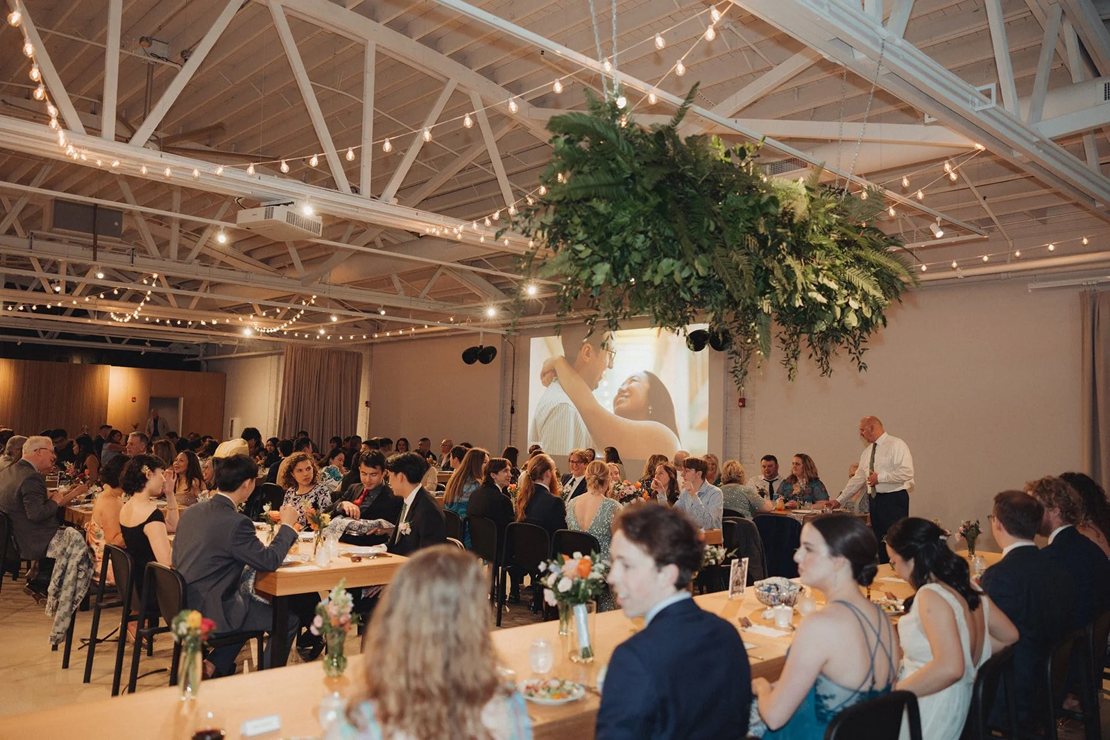 A large group of people in formal attire gathered in a decorated indoor space for a celebration, with a large screen displaying a couple embracing, and festive string lights hanging from the ceiling.