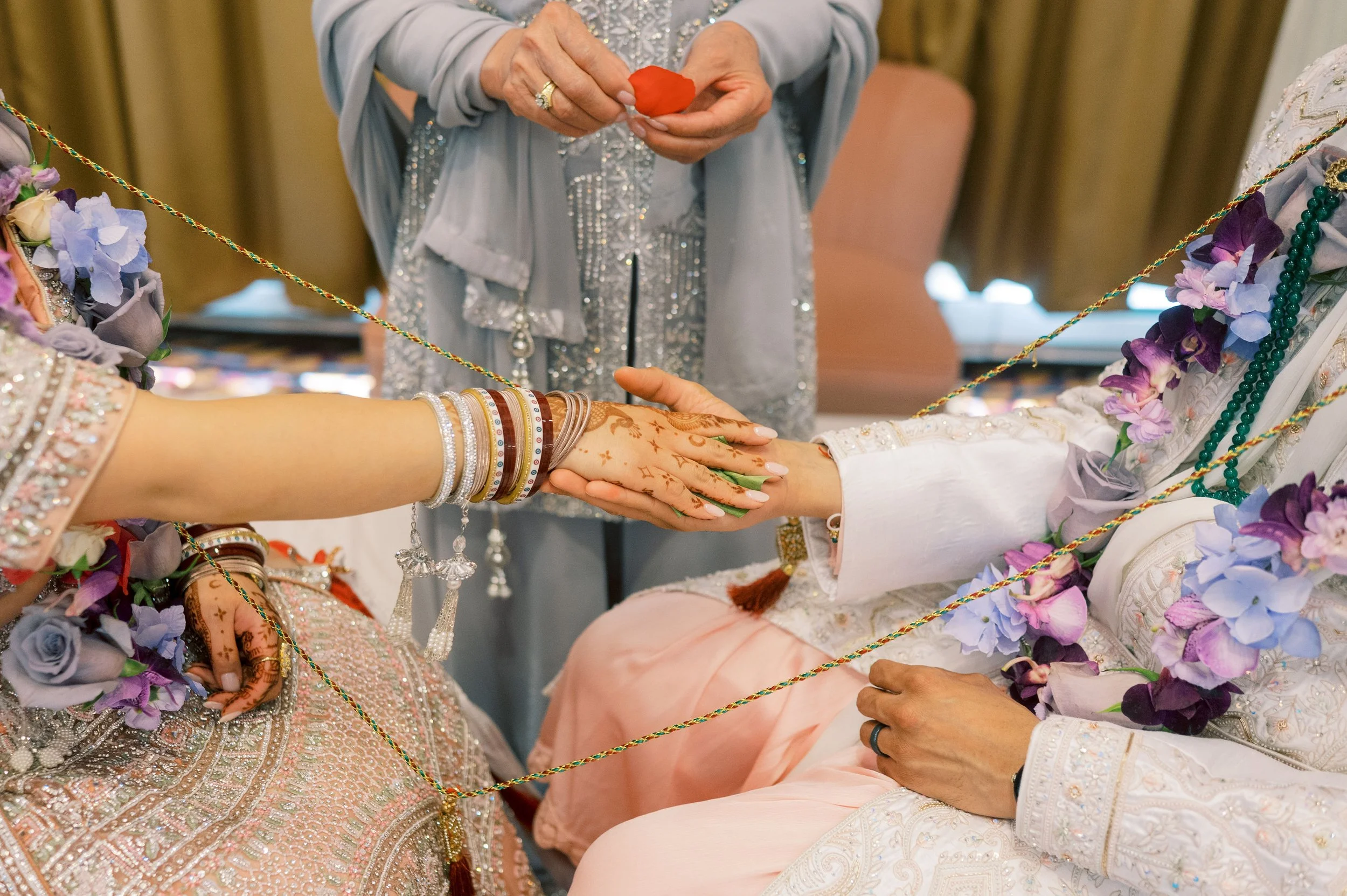 Close-up of a traditional Indian wedding ceremony where the bride and groom hold hands and exchange rings. The bride's arm is decorated with henna and jewelry, and the groom is dressed in ornate clothing. The scene is adorned with flowers and decorat