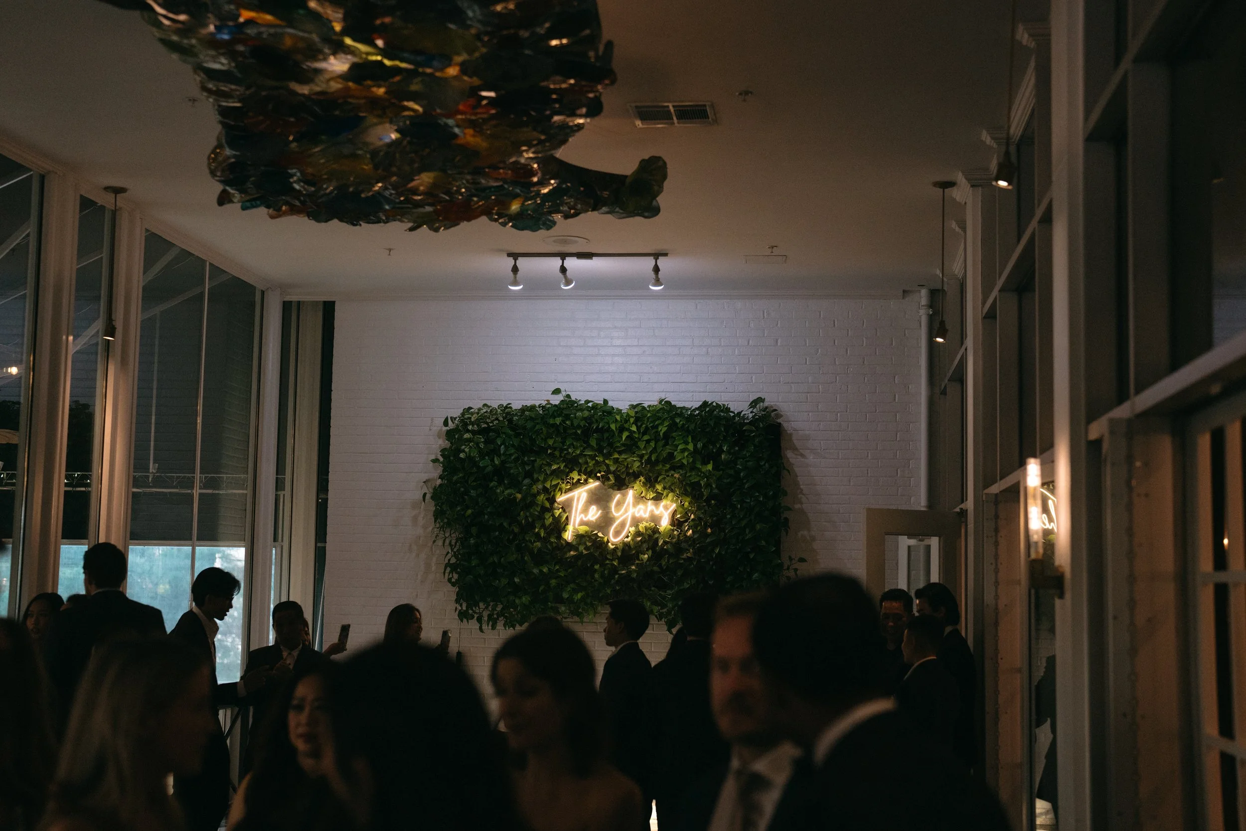 Nighttime indoor scene of a social gathering at a venue called 'The Yard,' with a white brick wall, green foliage, neon sign, and people dressed in formal attire.