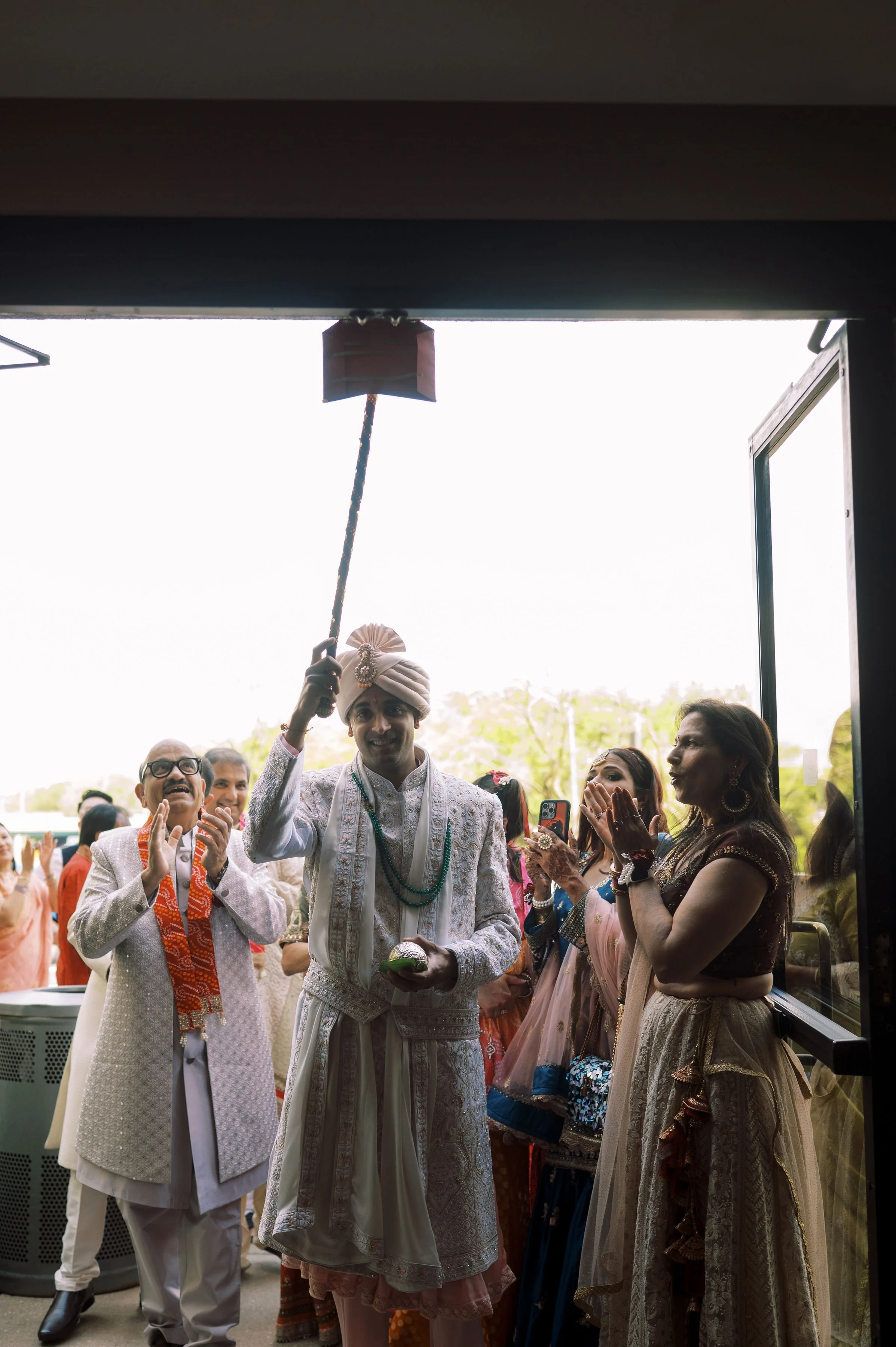 People dressed in traditional Indian attire, celebrating a wedding ceremony outside a building with an open door. The groom is holding a small object and smiling, while others around him claps and looks on.