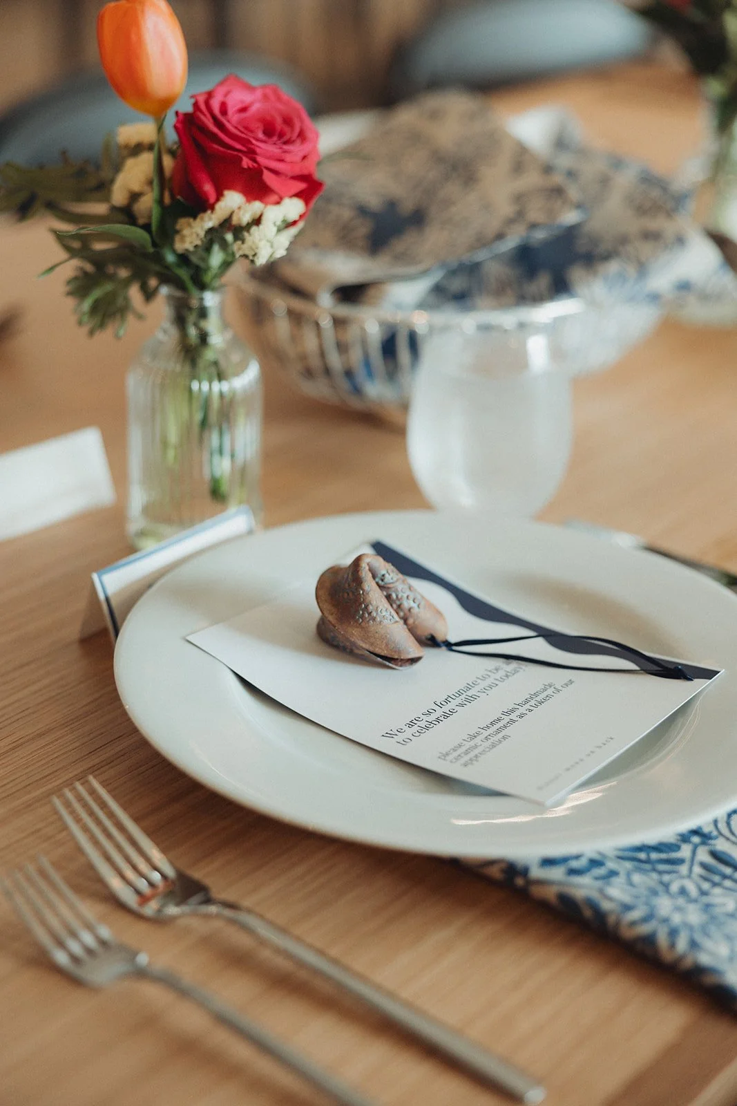 A table setting with a white plate, a menu, and a seashell necklace on top. In the background, there is a small vase with pink and orange flowers and a glass container with napkins. Silverware is placed on the wooden table.