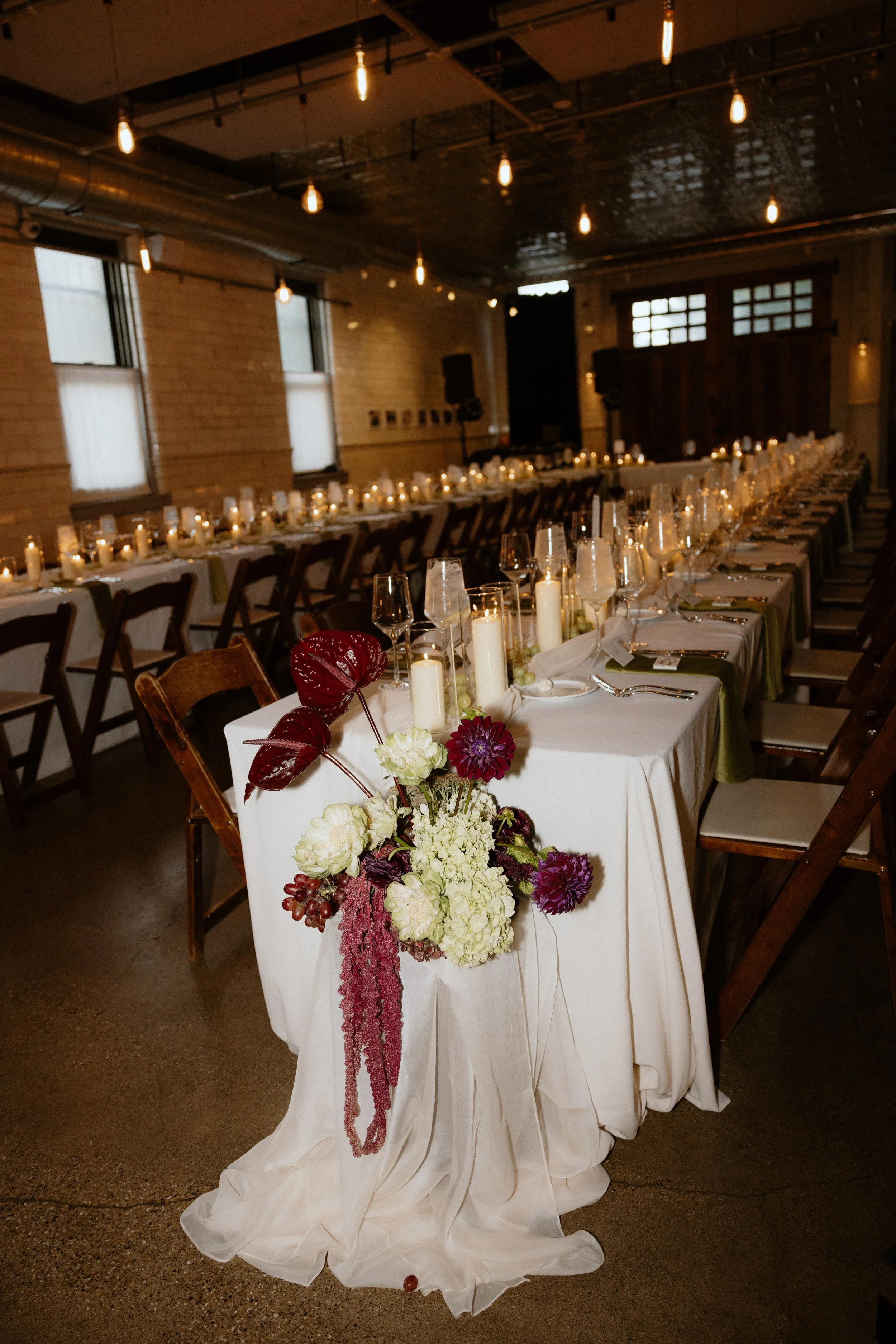 An elegant wedding reception table decorated with white tablecloth, candles, and a floral centerpiece with purple, white, and red flowers and greenery. The table is set with glassware and cutlery, and the room features exposed brick walls, wooden bea