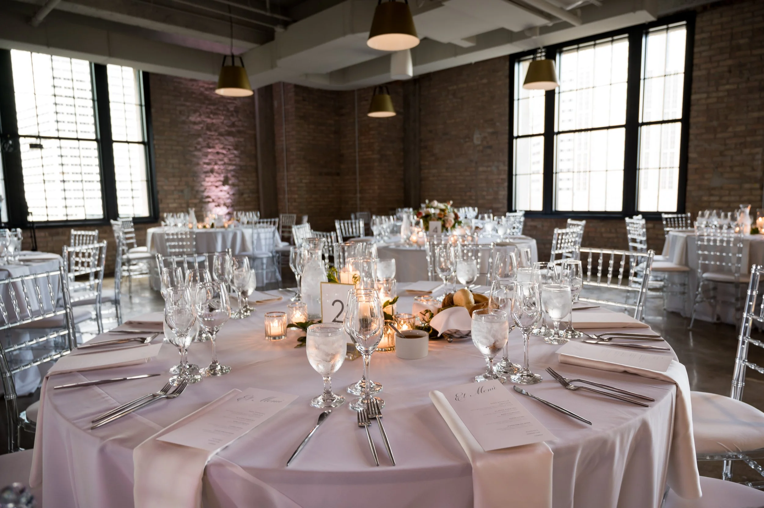 Elegant indoor event space decorated for a formal dinner, with round tables covered in white tablecloths, set with glassware, cutlery, and menus, featuring centerpieces of flowers and candles. Large windows and exposed brick walls dominate the room.