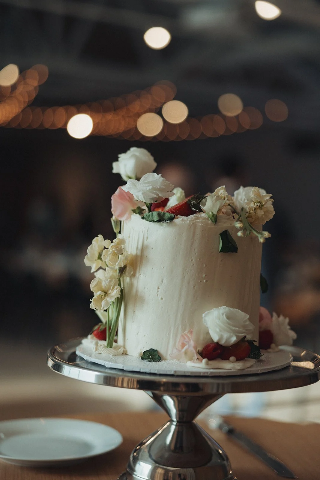 A tall white cake decorated with fresh flowers and strawberries on a silver cake stand at a celebration or wedding reception.