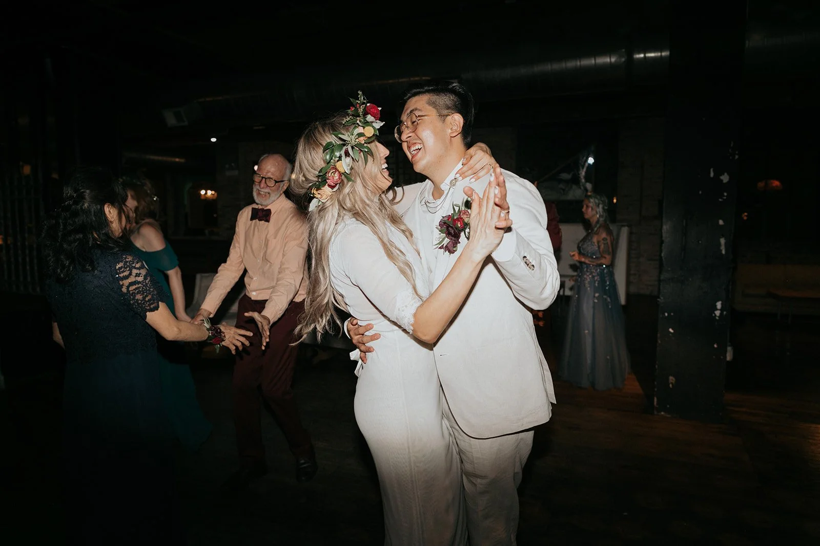 A couple dressed in wedding attire dance closely, smiling at each other, with friends in the background at a celebration.