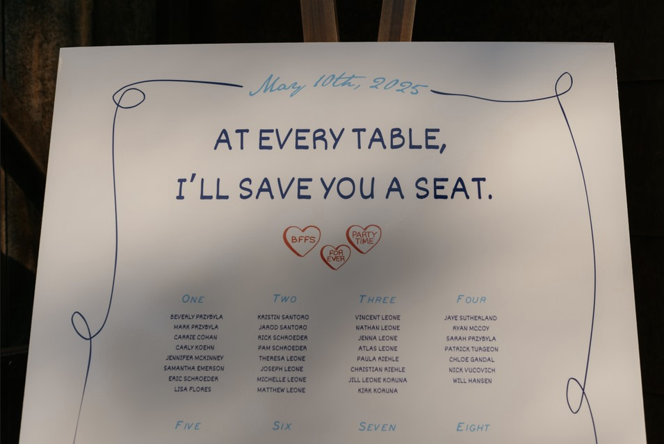 A white wedding or event sign with blue and red text. The top of the sign has the date "May 10th, 2025" written in cursive. The main message says, "At every table, I'll save you a seat." Below are numbered lists of names under headings for tables one