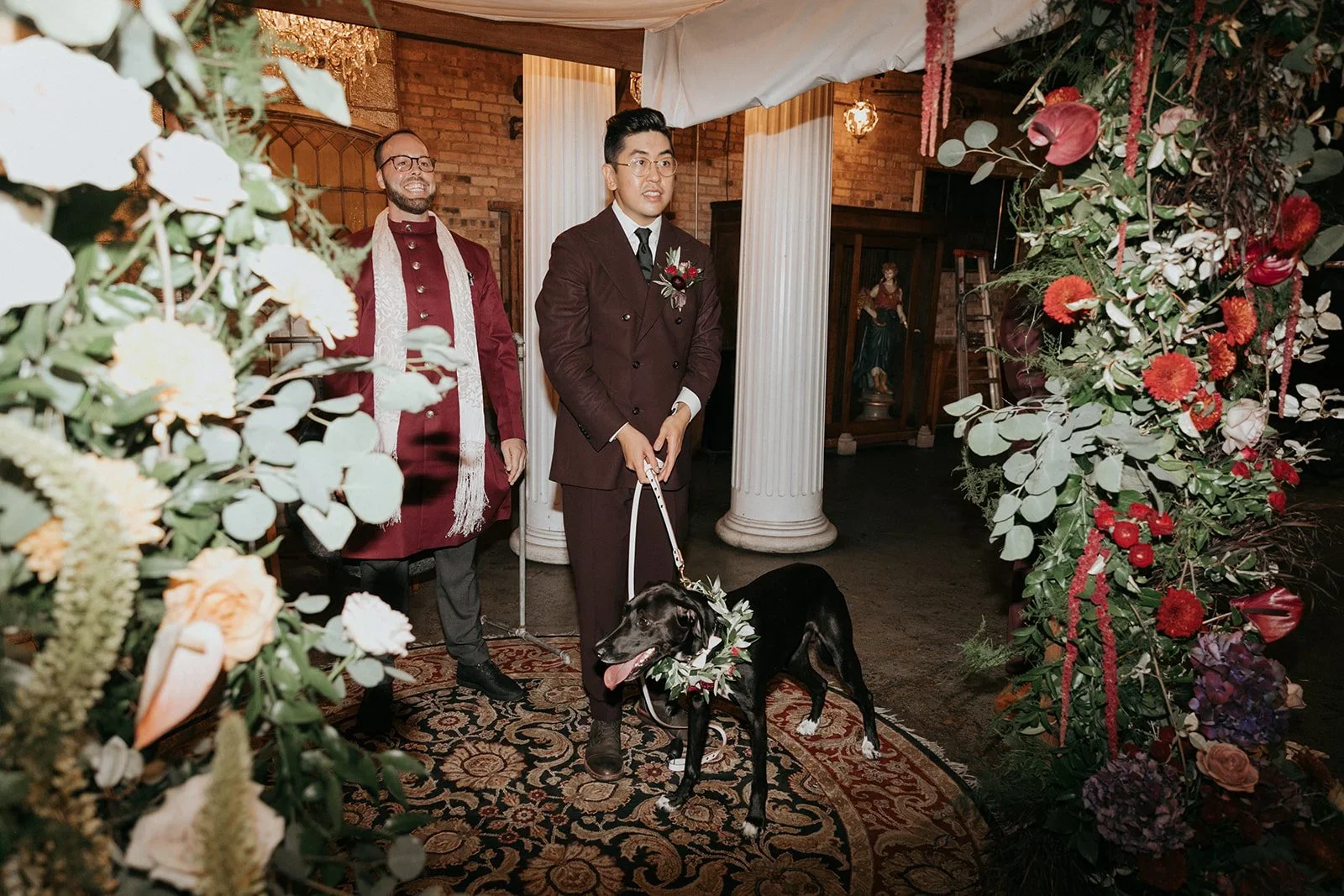 A same-sex wedding ceremony indoors, with two men standing, one dressed in a dark suit with a leash, holding a black dog with a floral collar, in front of floral and greenery decorations, with stone columns and a brick wall in the background.