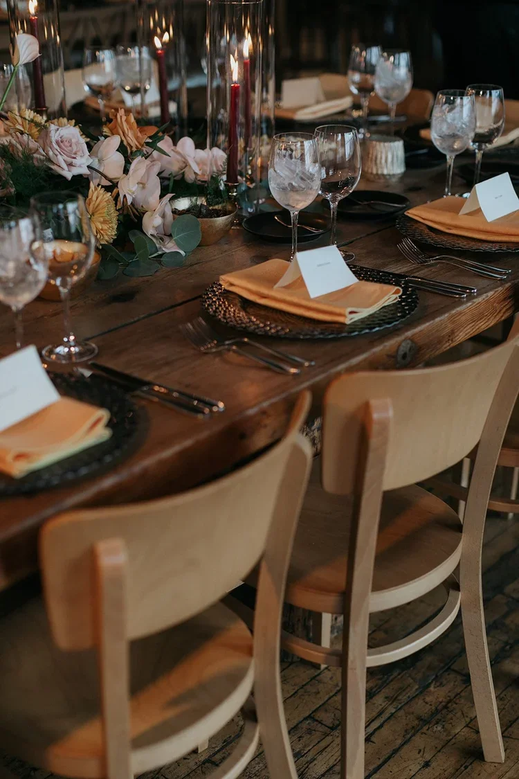 Elegant dining table set with black plates, gold napkins, wine glasses, and floral centerpiece with pink roses and greenery.