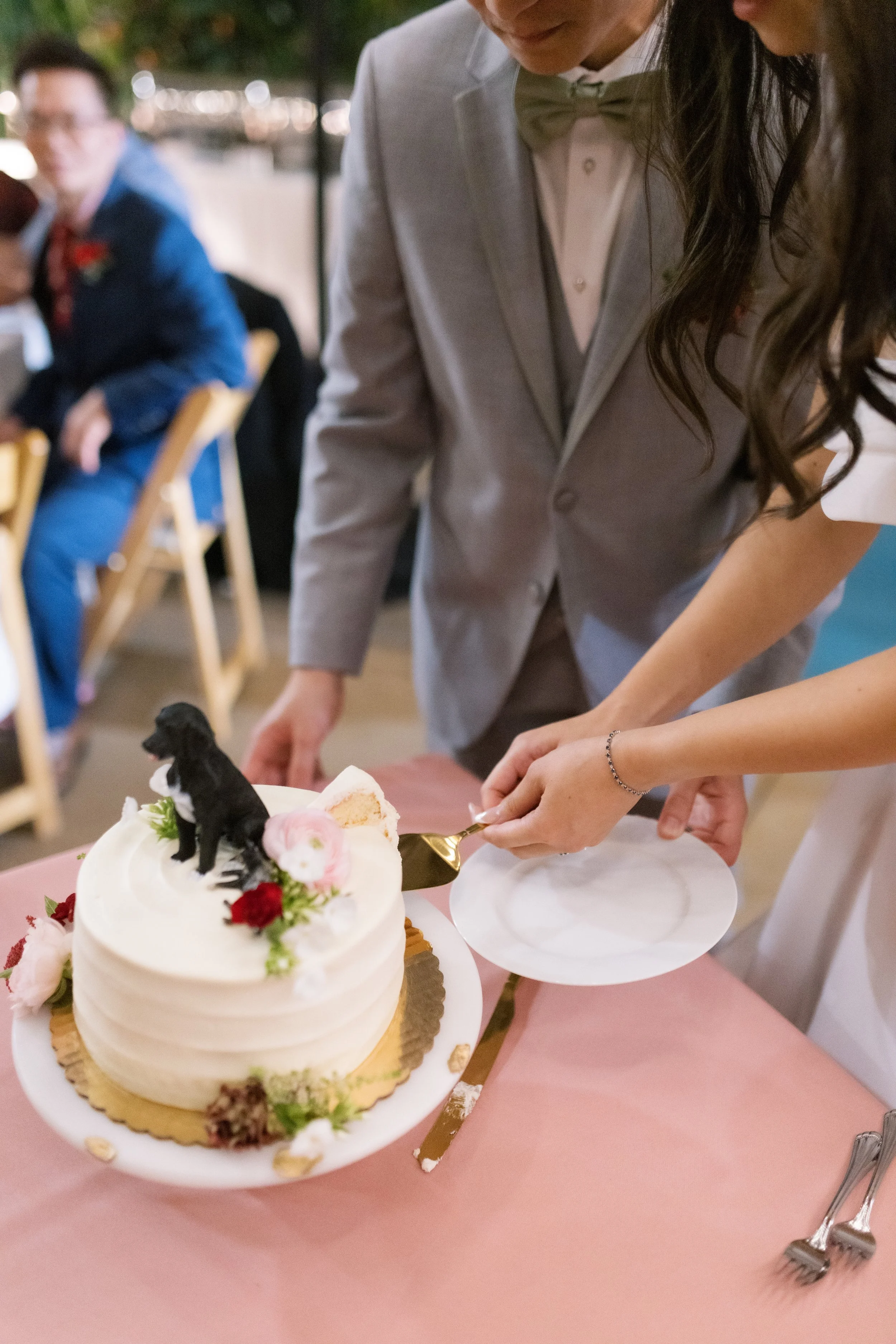 People at a celebration cutting and serving a layer cake decorated with flowers and a dog figurine, with a pink tablecloth, forks, and plates visible.