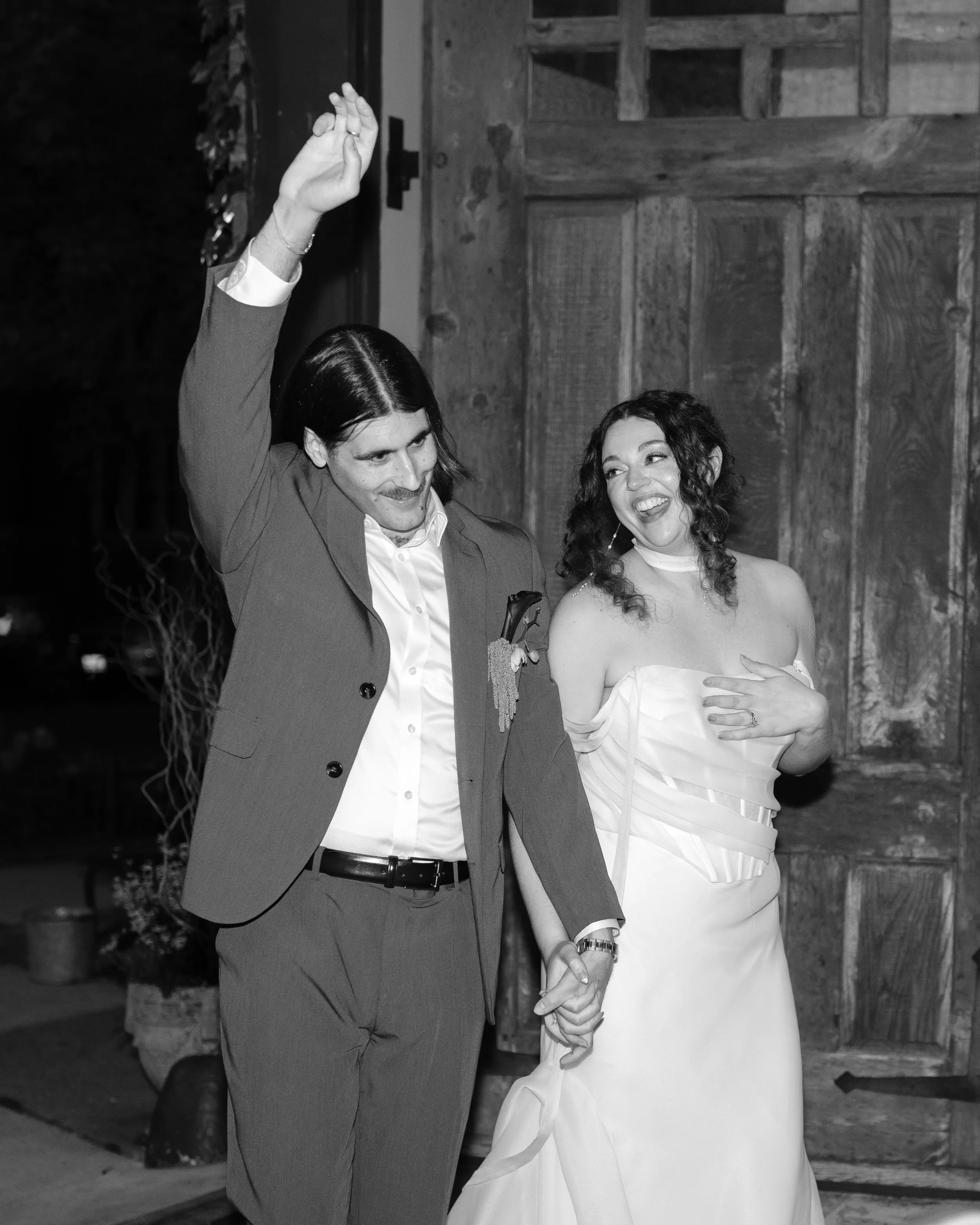 A man in a suit and a woman in a strapless dress holding hands and smiling at an event, possibly a wedding, with a wooden wall in the background.