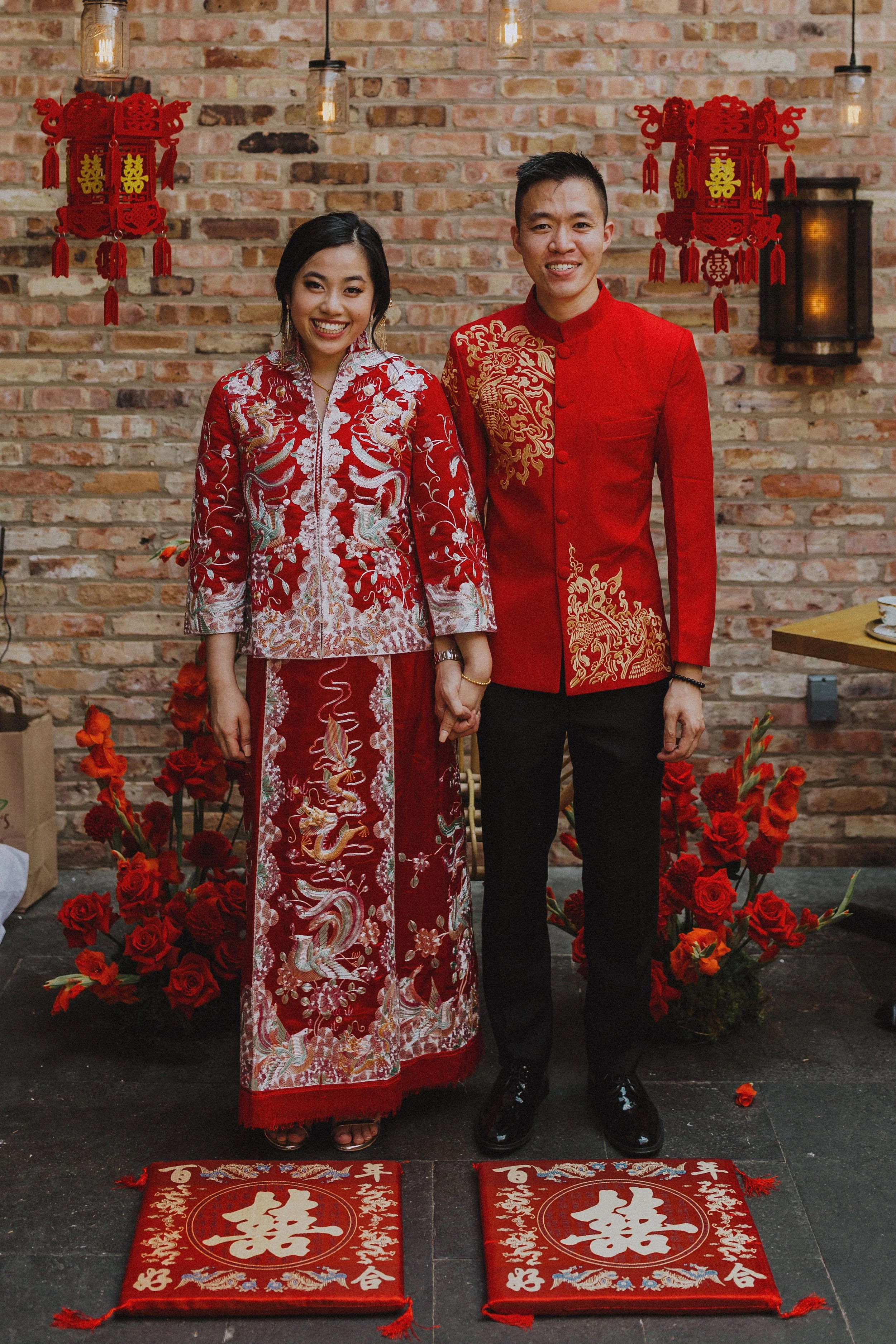A couple dressed in traditional Chinese wedding attire standing together, holding hands, surrounded by red roses and Chinese decorations, on a black floor with red Chinese wedding cushions with white Chinese characters.