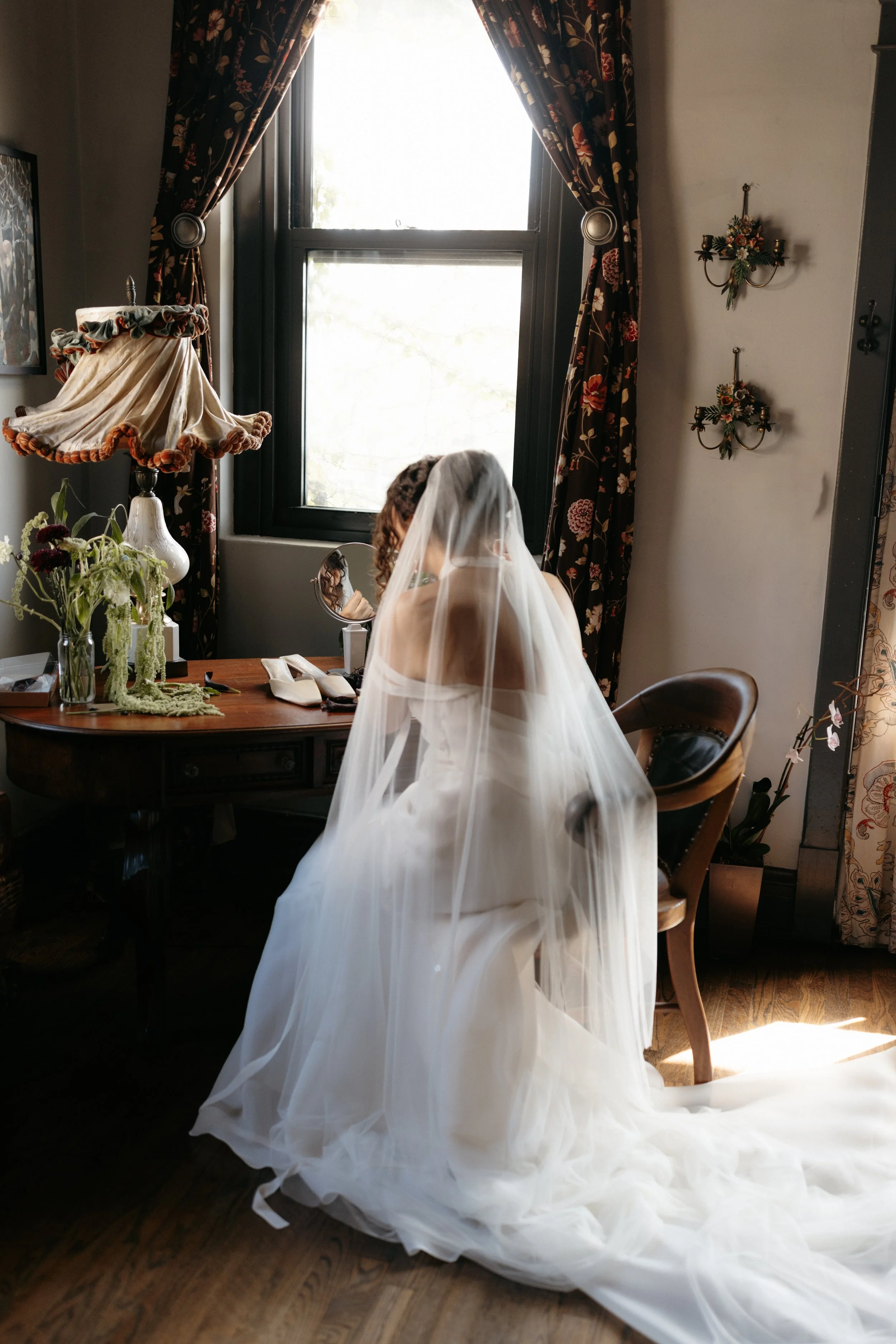 A bride in a wedding dress with a veil sitting at a vanity table in a room with floral curtains, a vintage lamp, and wall-mounted candle holders with flowers.