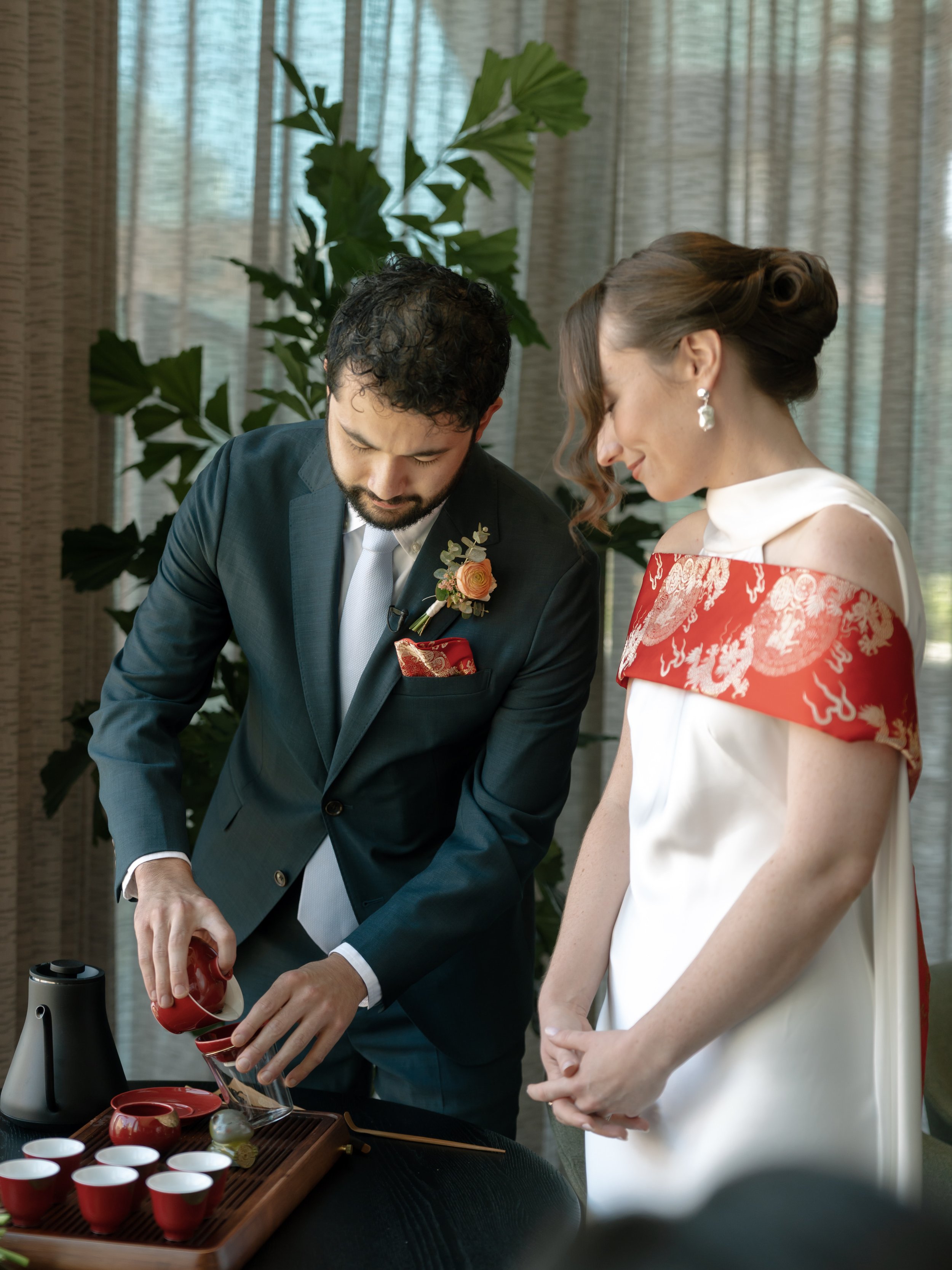 A couple dressed in formal attire participating in a traditional Japanese tea ceremony. The man is pouring tea from a red teapot into small red cups on a tray, while the woman watches with a gentle smile. The setting includes a large leafy plant and 