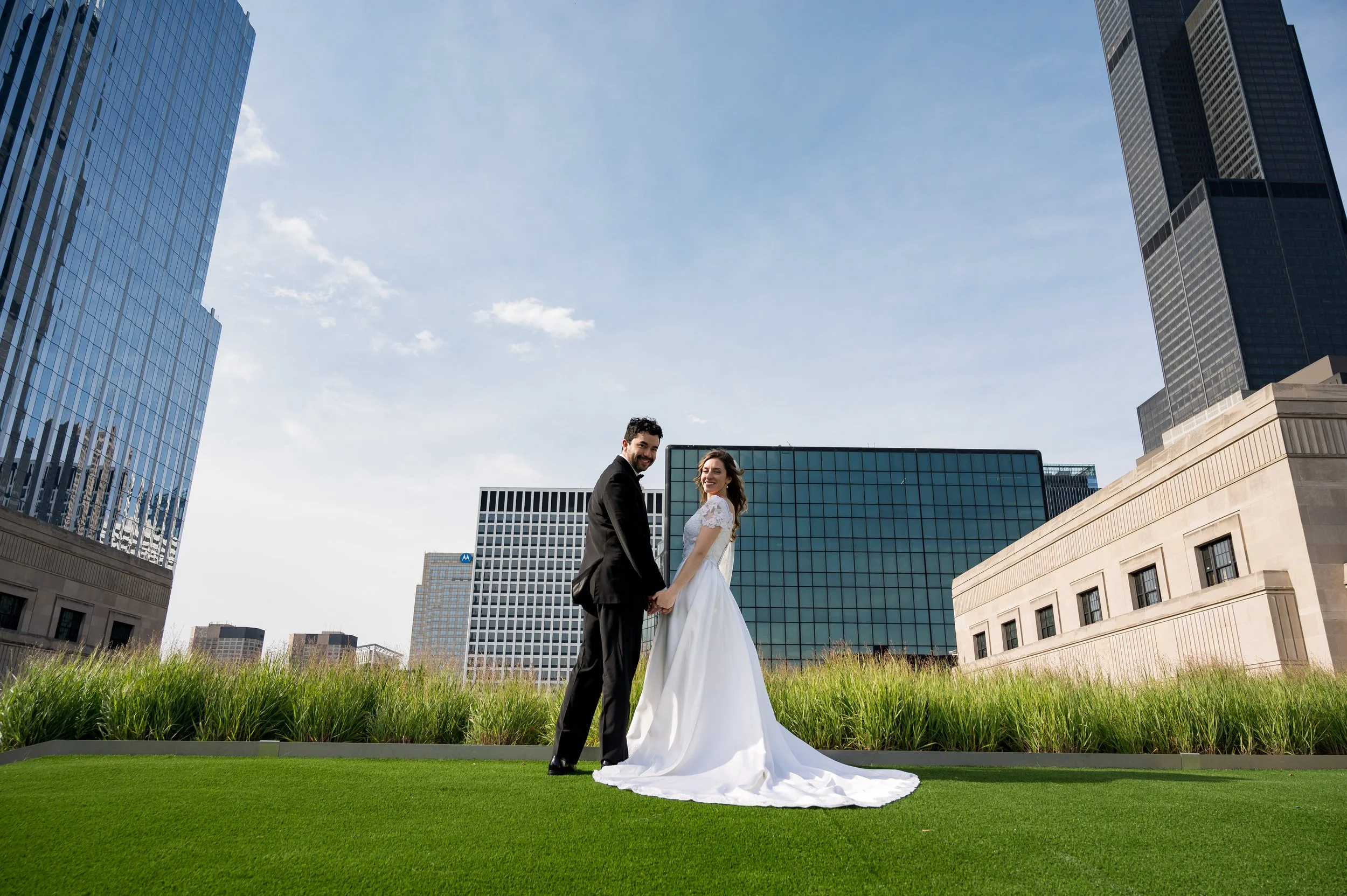 A bride and groom holding hands and smiling in an urban setting with tall modern glass buildings behind them. The bride is wearing a white wedding dress and the groom is in a black suit. They are standing on a grassy area under a blue sky.