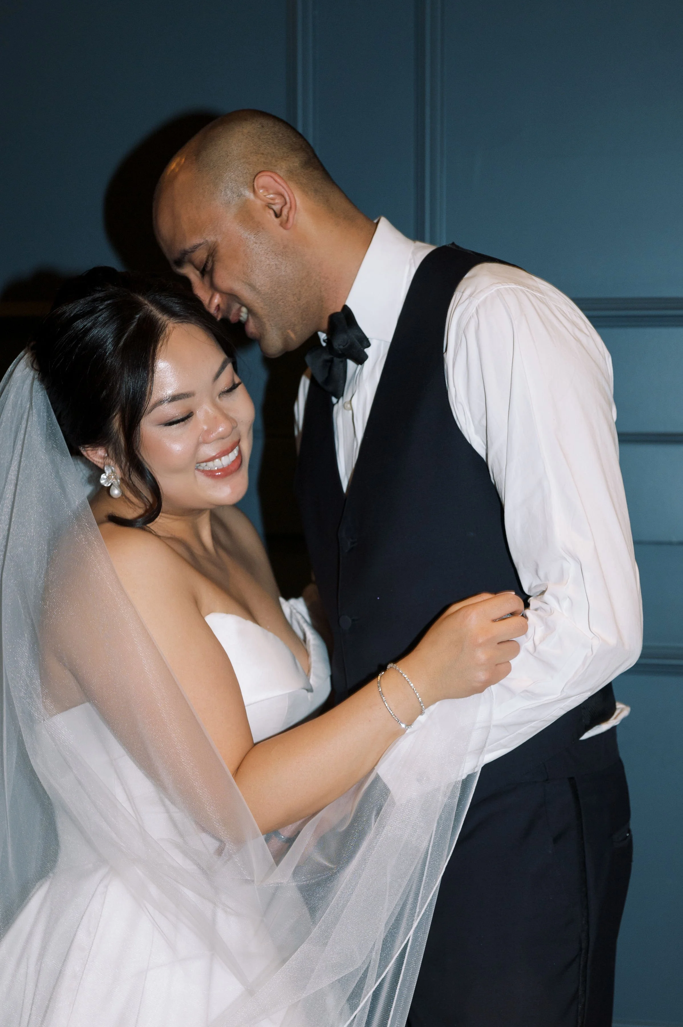 A bride and groom embrace each other at their wedding, smiling and looking happy, with the bride wearing a white wedding dress and veil, and the groom in a tuxedo with a bow tie, standing against a blue wall.