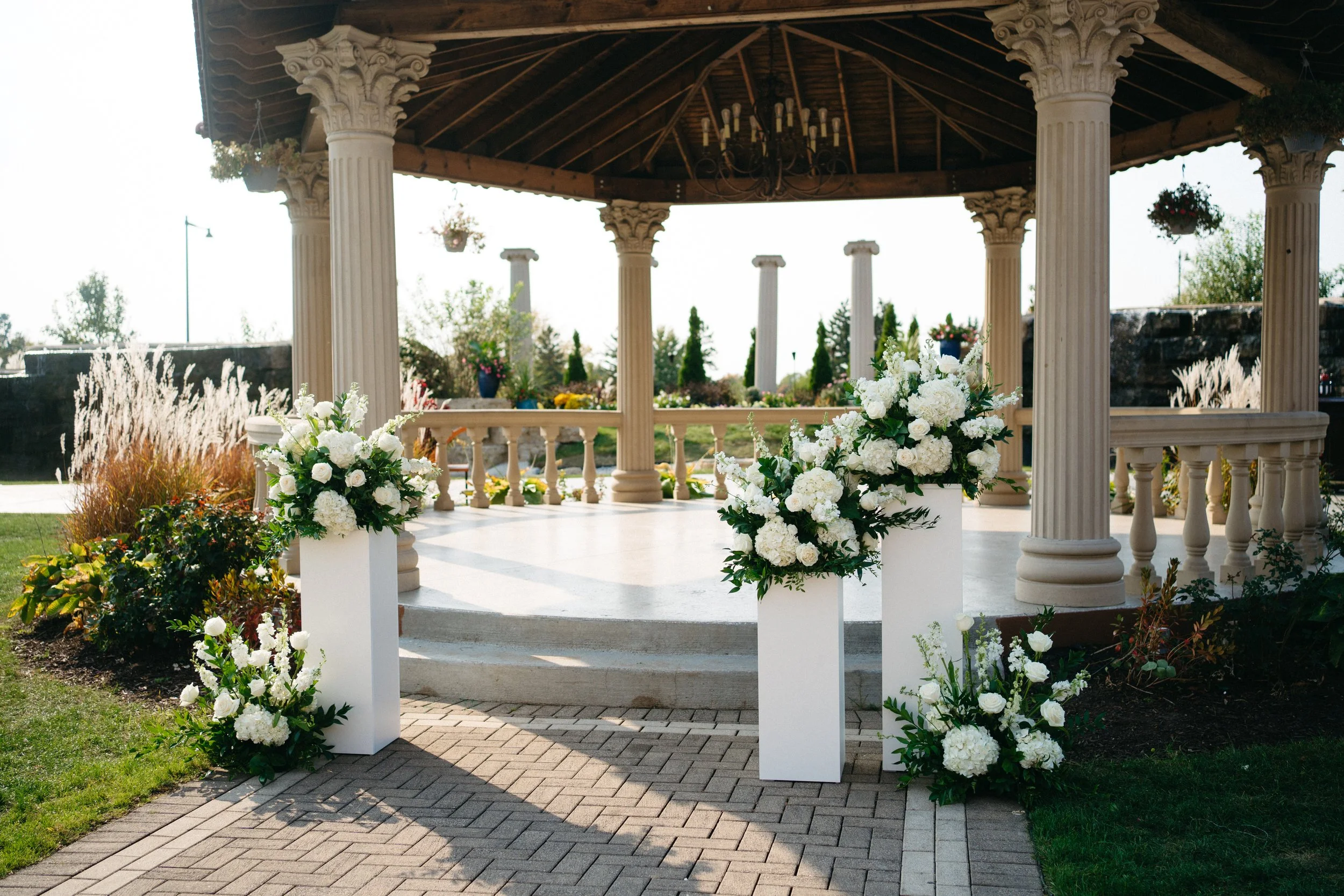 A gazebo with six cream-colored columns, a chandelier, and hanging flower pots. The gazebo is decorated with large white floral arrangements and is surrounded by a garden with various plants, flowers, and trees.