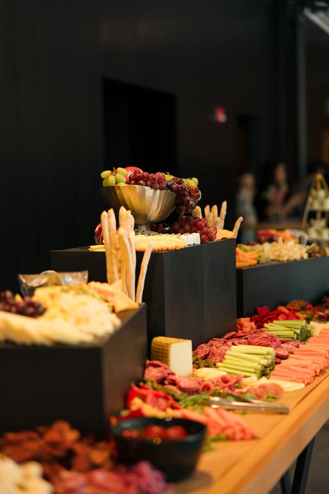 Variety of fruits, cheeses, and charcuterie on a buffet table with black display blocks and a dark background.