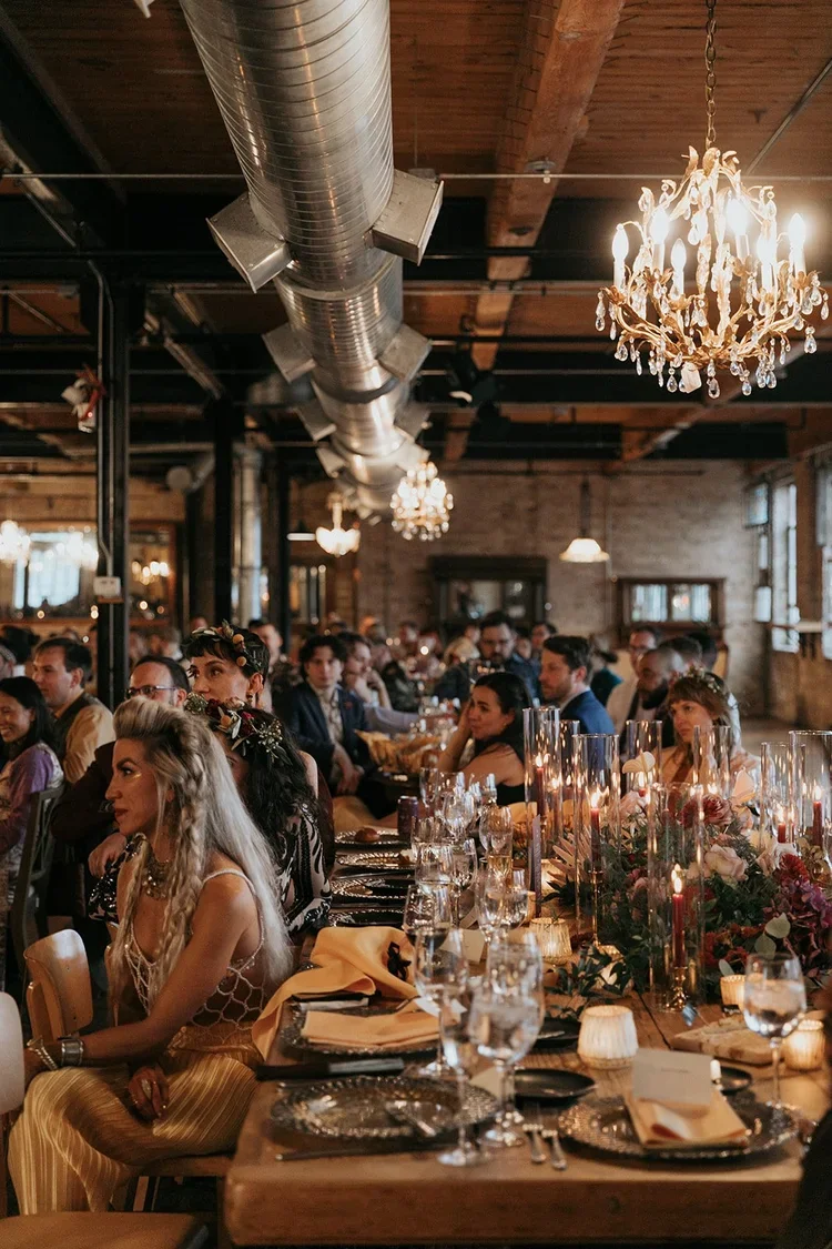 People dining at a long table decorated with flowers and candles in a rustic restaurant with exposed brick walls, wooden beams, and chandeliers.