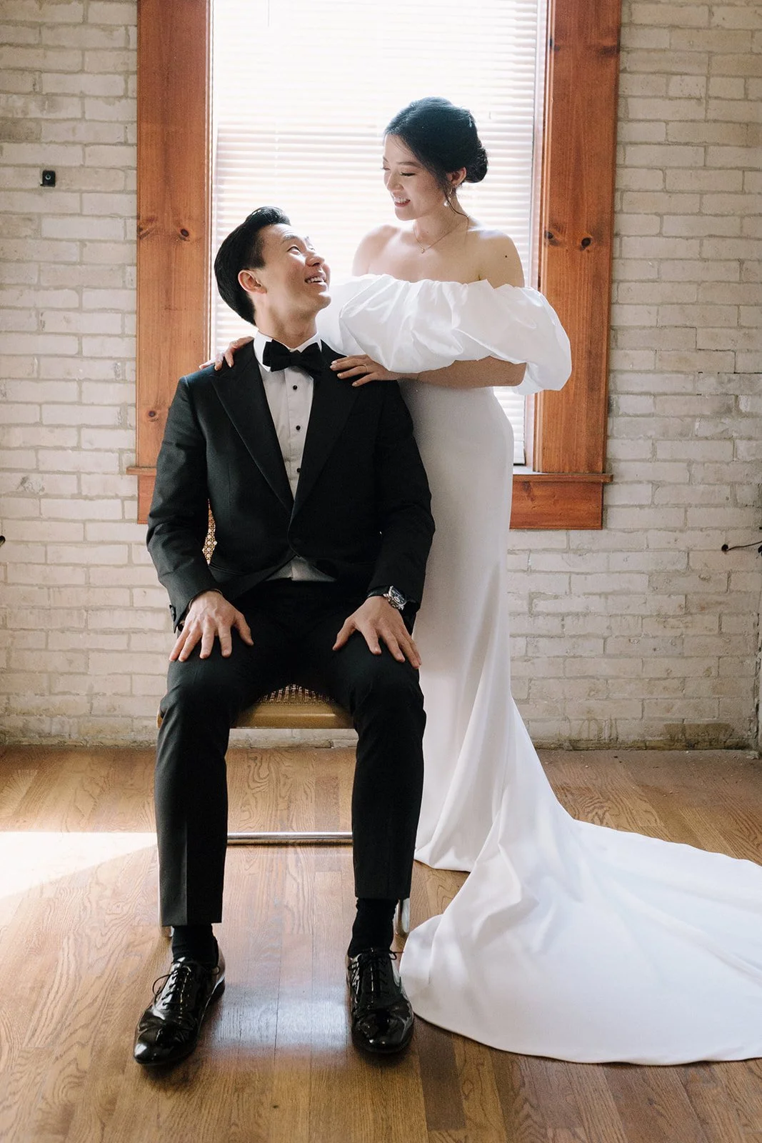 A bride and groom in wedding attire inside a room with a brick wall and wooden window frame. The groom is sitting on a chair, wearing a tuxedo, and the bride is standing behind him, touching his shoulders and smiling while looking at him.