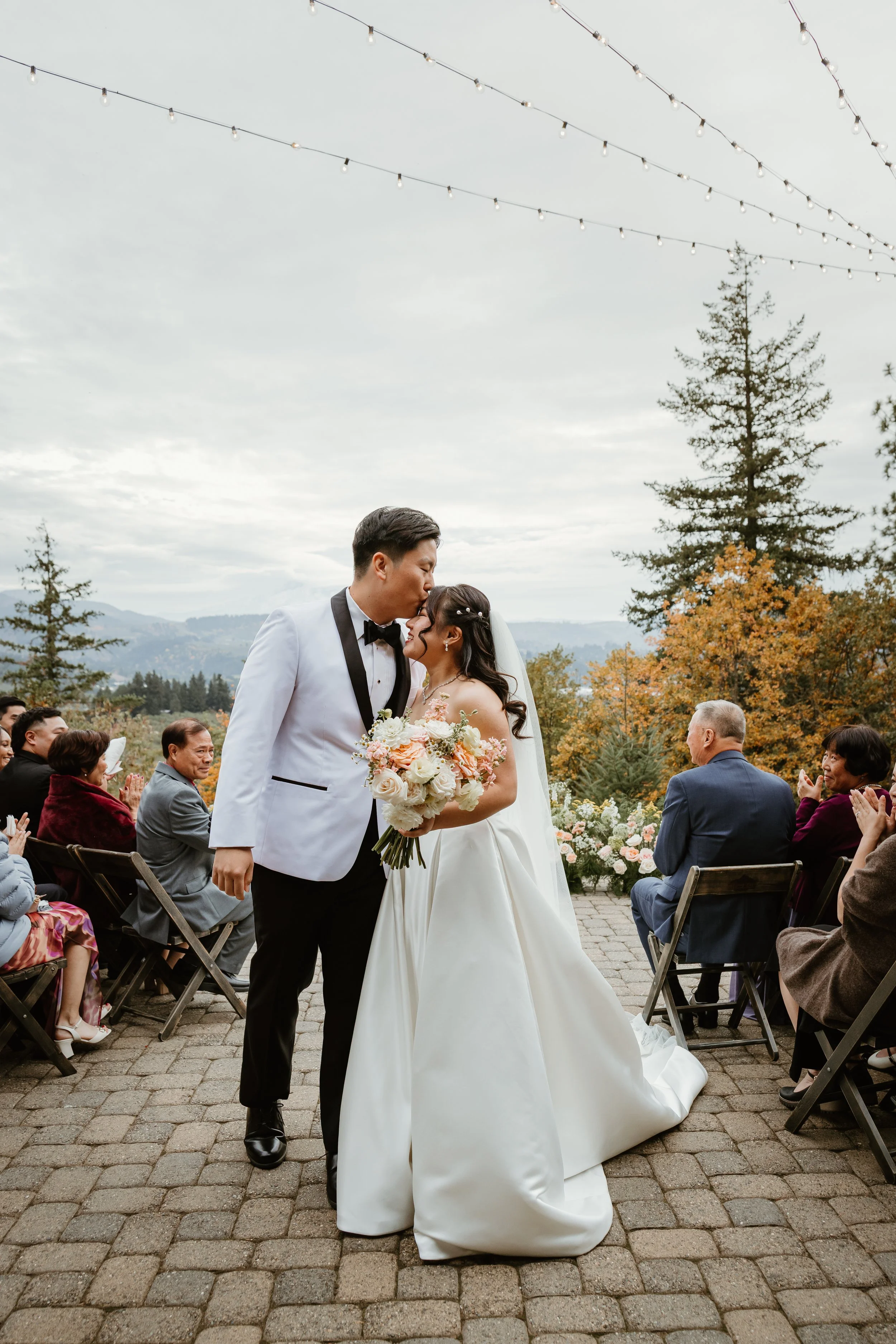A newlywed couple kissing at their outdoor wedding ceremony, surrounded by seated guests, with trees and mountains in the background, and string lights overhead.