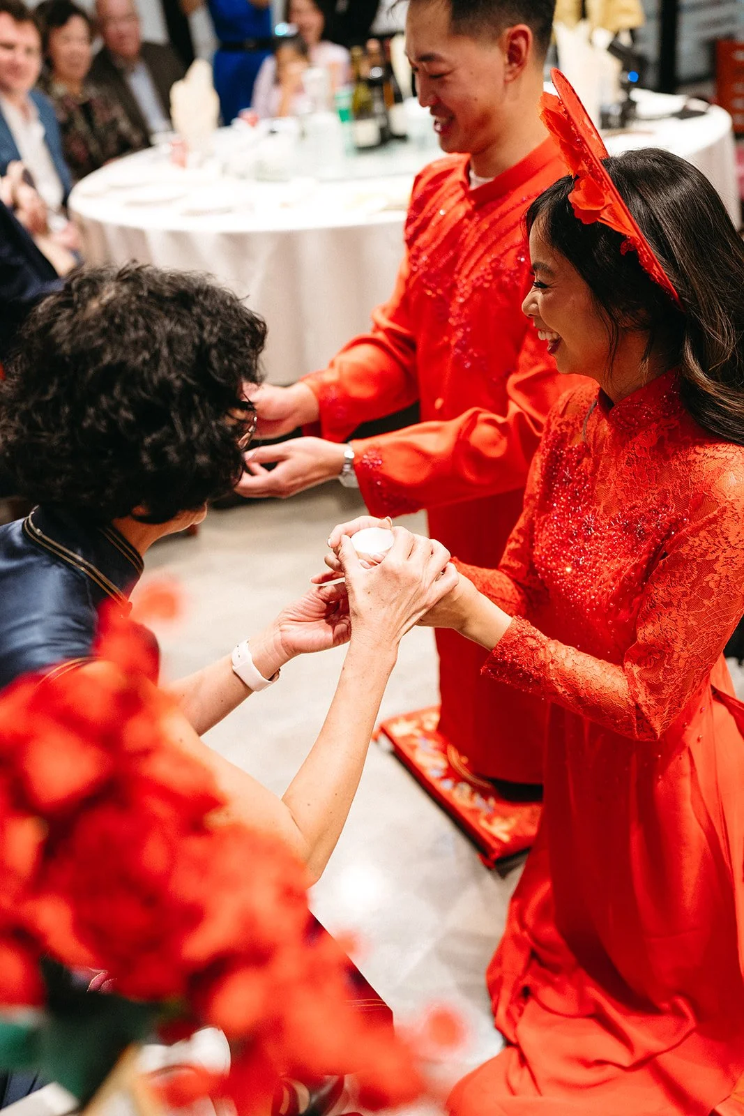 A traditional Chinese tea ceremony during a wedding, with a bride and groom in red dresses offering tea to an elder, surrounded by family and friends.