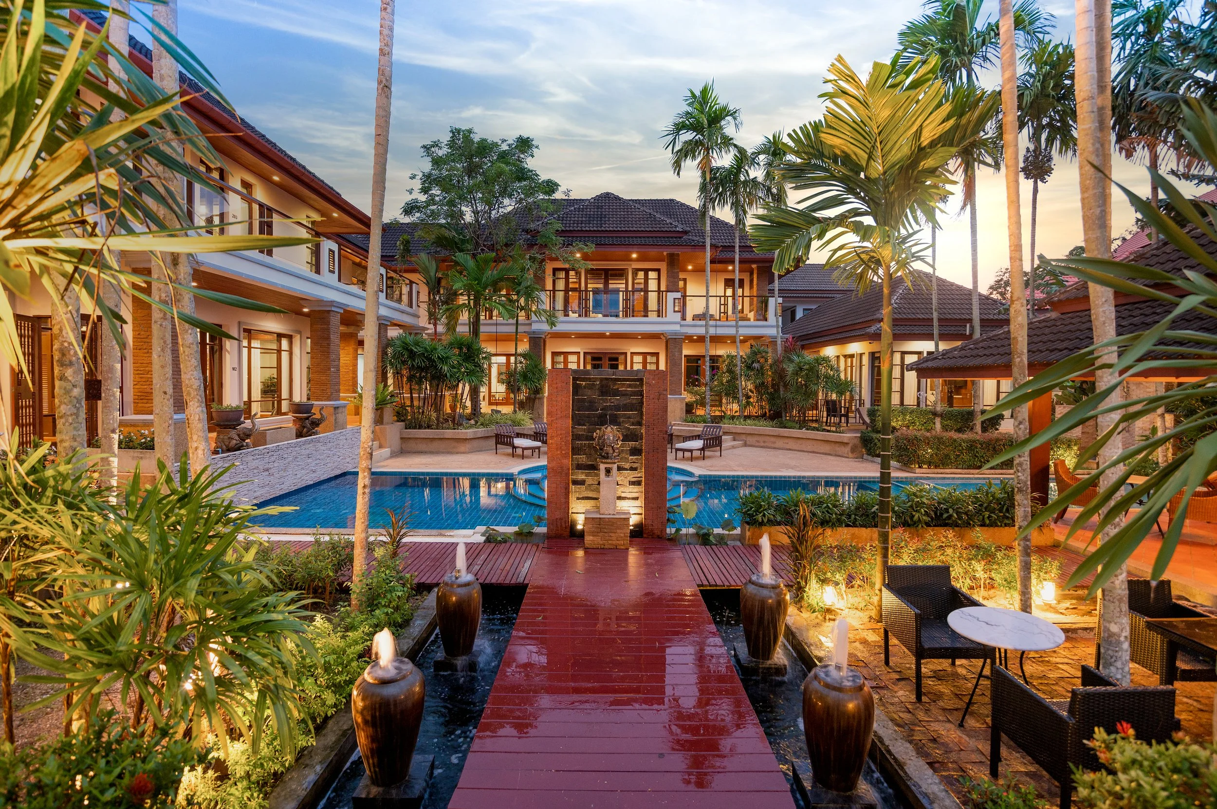 Luxury resort courtyard with a swimming pool, tropical plants, a red walkway, and two-story buildings, at sunset.