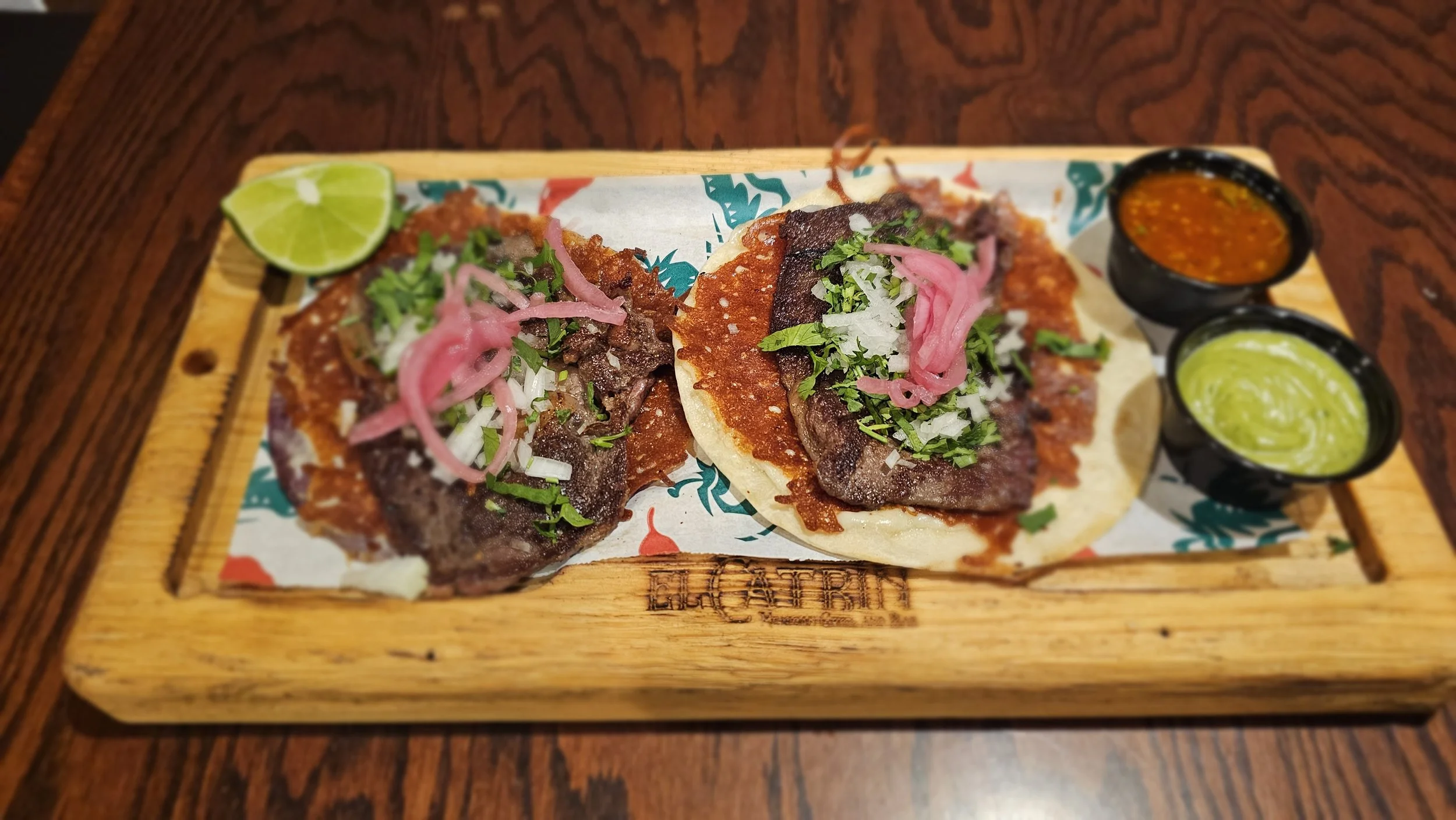 Two Mexican street tacos with grilled meat, topped with chopped onions, cilantro, and pickled pink onions, served on a wooden platter with lime wedges and two small bowls of salsa, one red and one green.