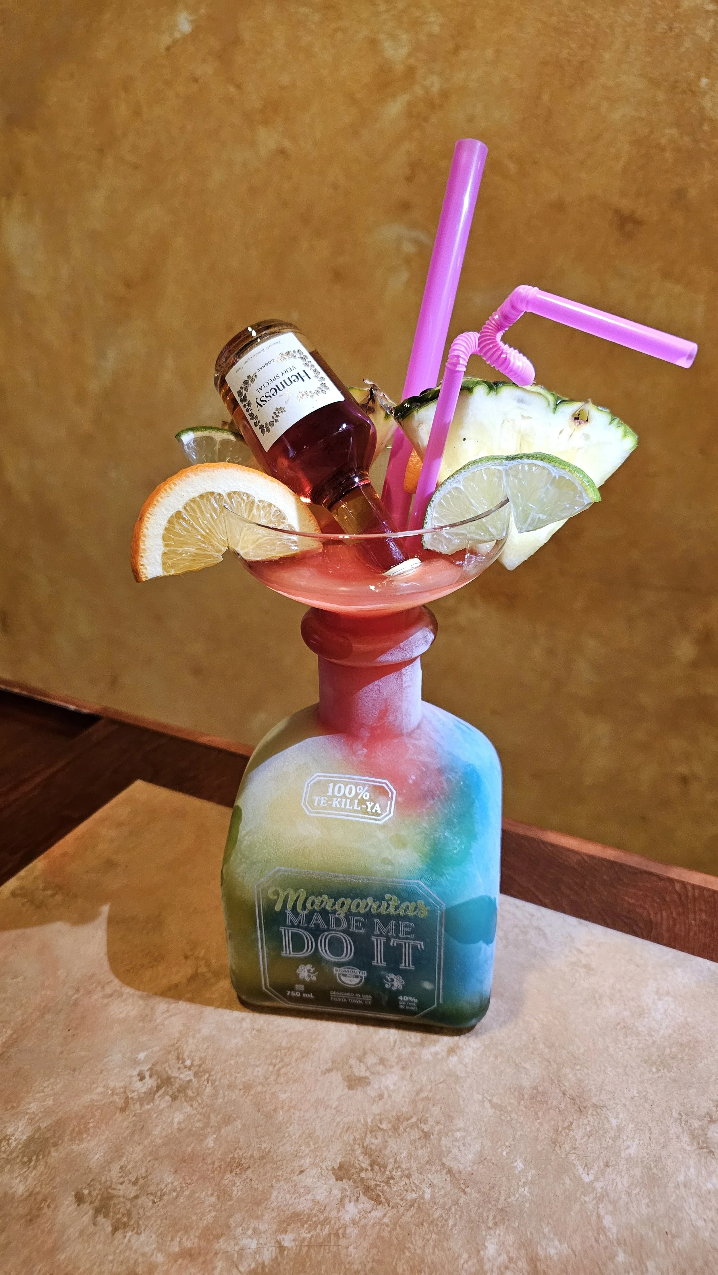 A humorous cocktail glass with various fruit slices, a small bottle of Cherry Hennessy, and colorful straws, all placed on a repurposed bottle with a playful label.