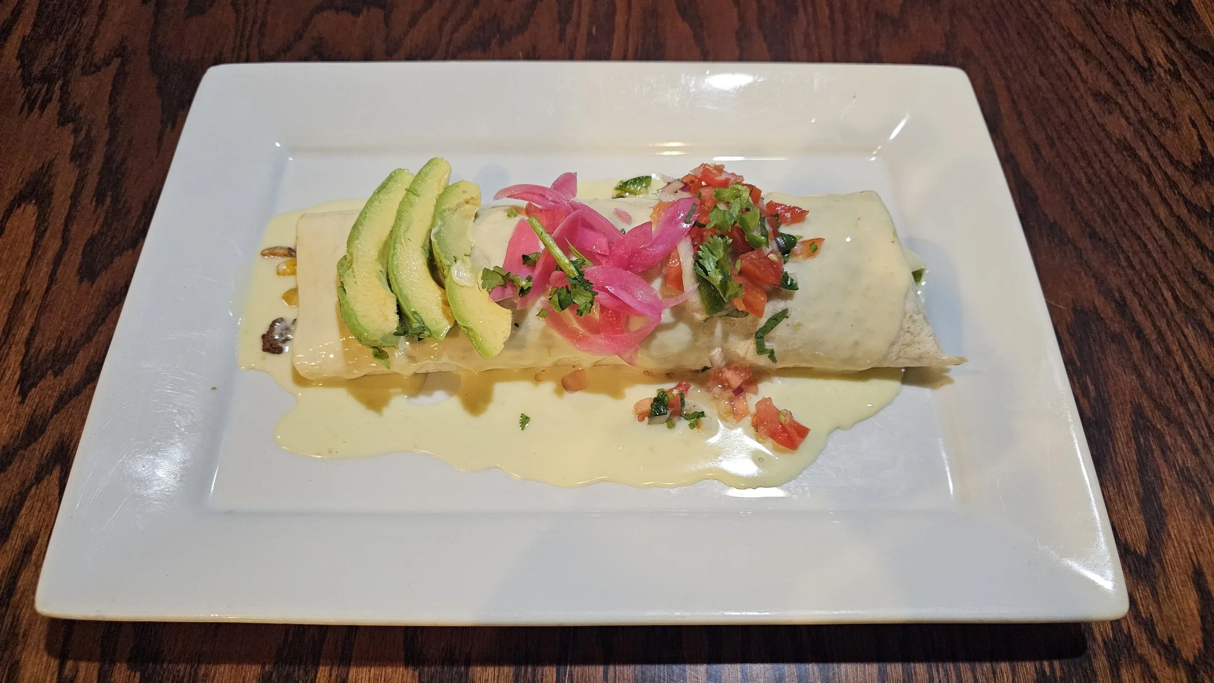 Plate of Mexican food with sliced avocado, pickled red onion, and chopped tomato, served with creamy sauce on a white square plate.