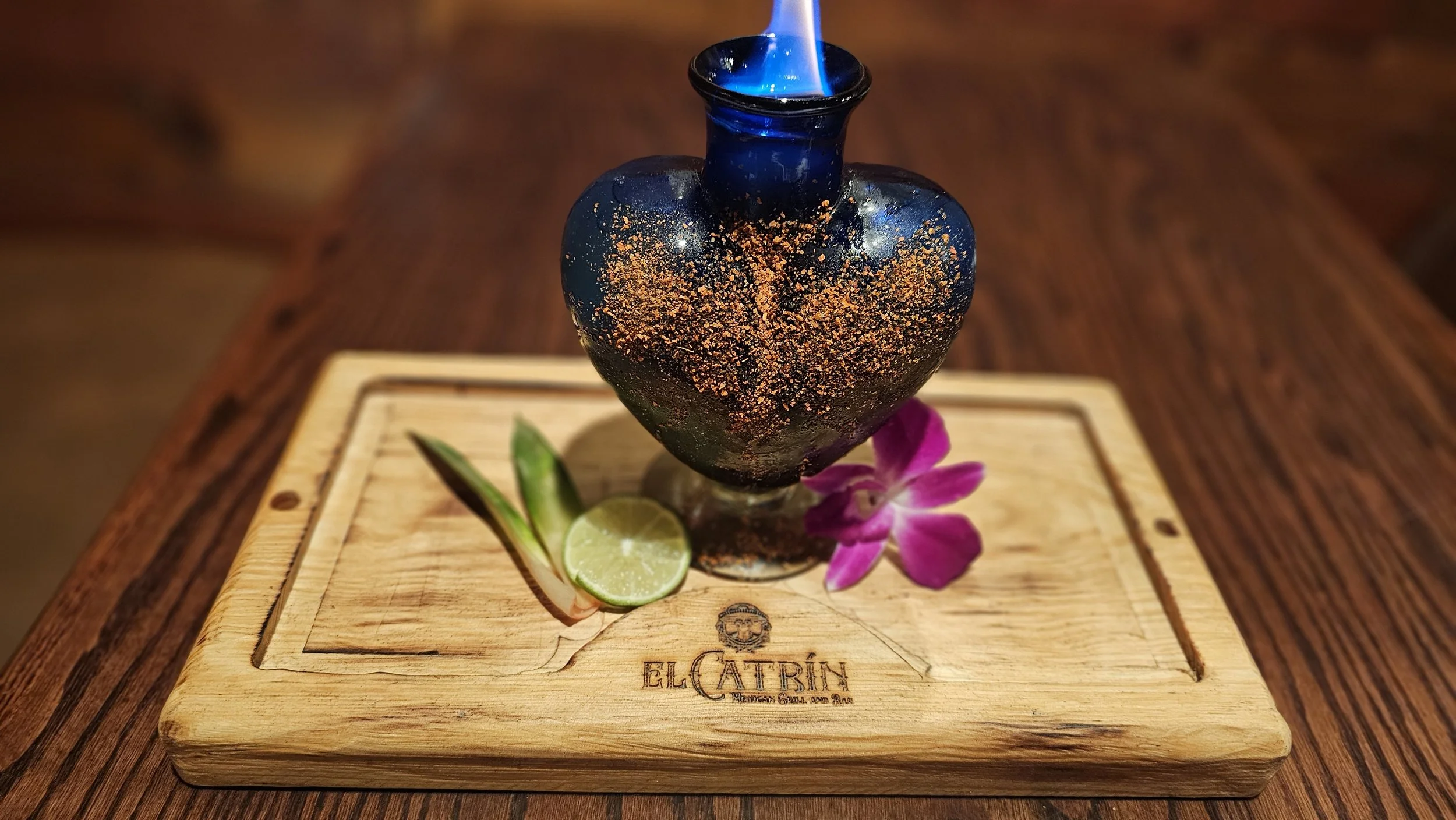 A blue torch with a flame burning inside a heart-shaped glass vessel, placed on a wooden serving board with slices of lime, a green leaf, and a purple flower, with the words "El Catrin" branded on the board.