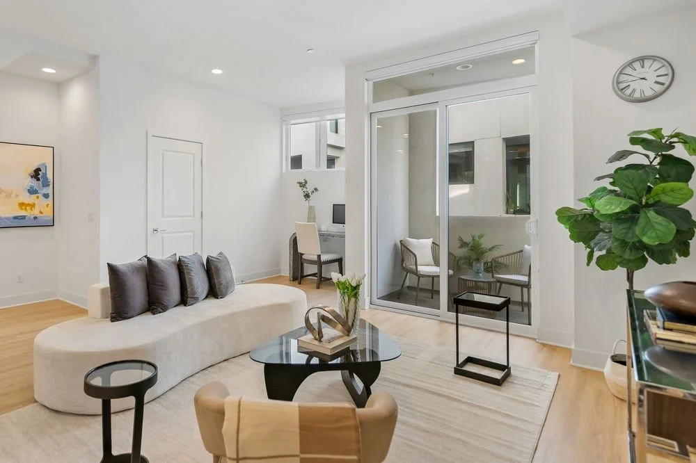 Modern living room with white walls, wooden flooring, a beige curved sofa with dark cushions, a glass coffee table with a sculpture and flowers, a large indoor green plant, and a small patio with outdoor seating visible through sliding glass doors.