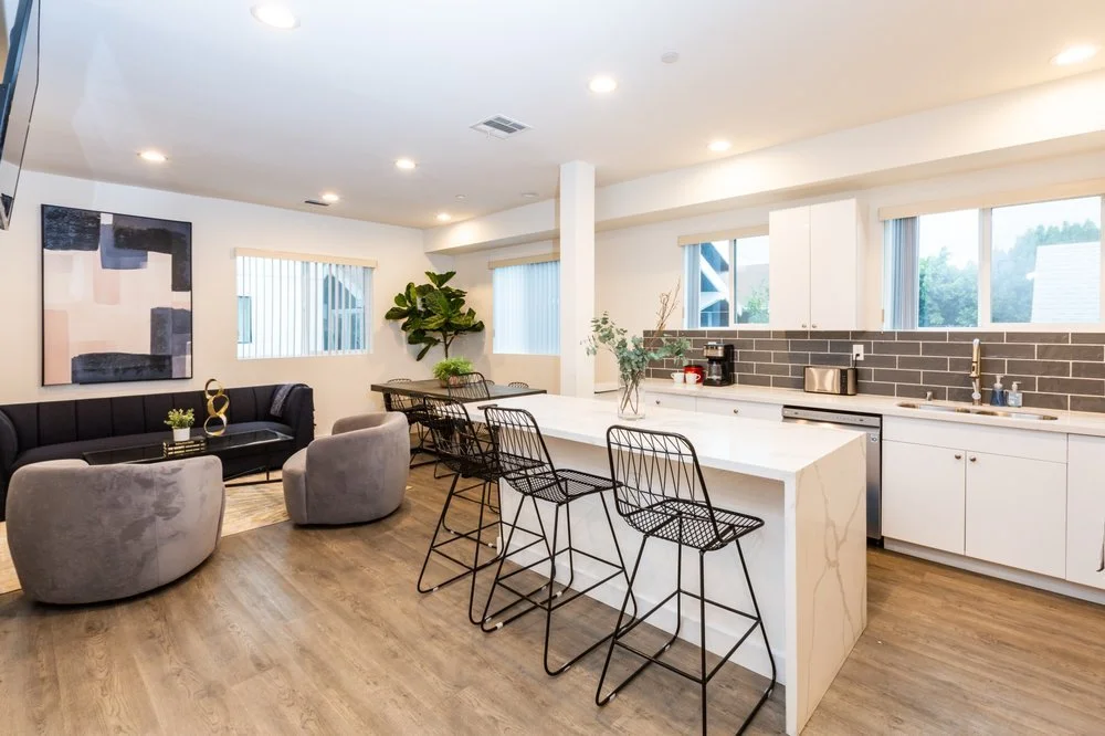 Open-concept living room and kitchen with modern decor, black couch, grey chairs, white kitchen island, and large windows.