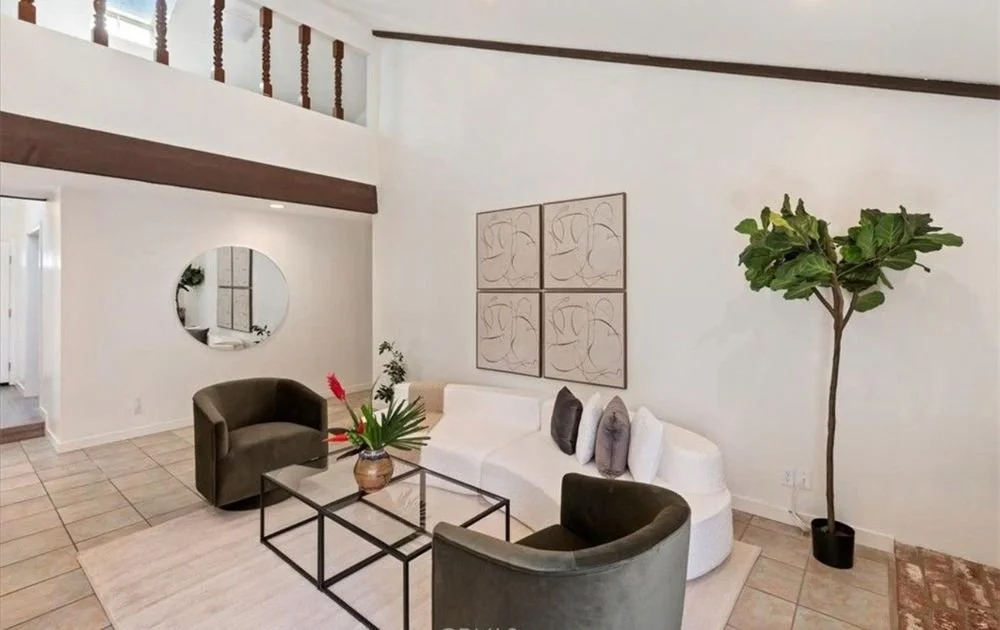 Modern living room with white walls, a white curved sofa, two dark green armchairs, a rectangular glass coffee table, a decorative plant in a vase, and a large potted plant in the corner. There are four abstract art pieces on the wall and a round mir