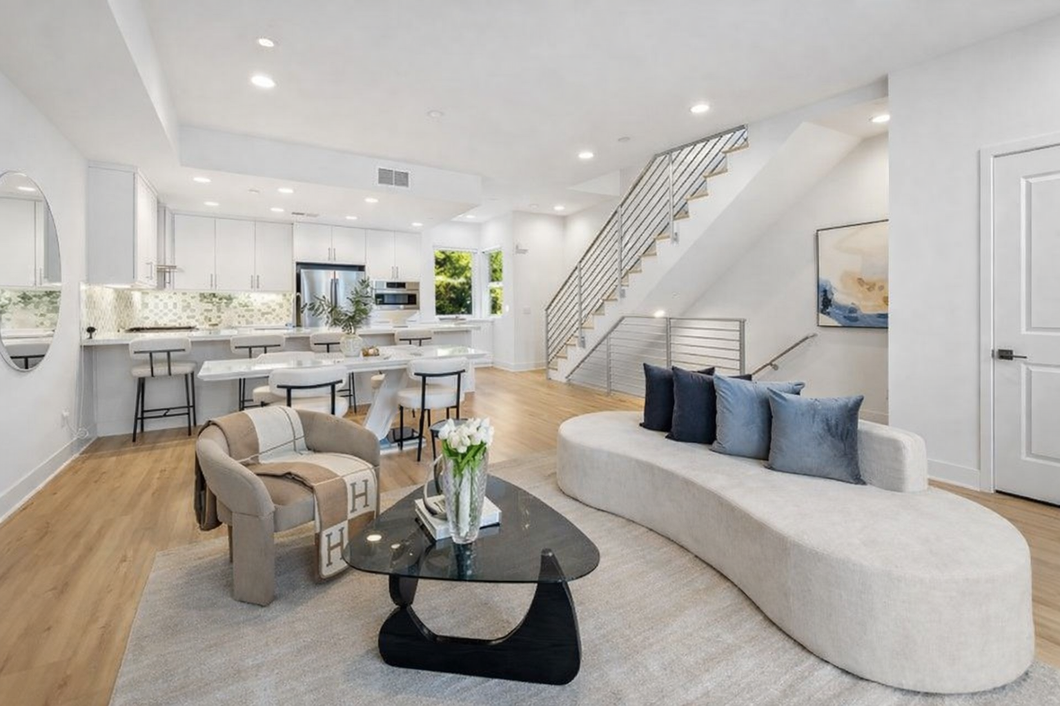Open-concept living room and kitchen with modern decor, light-colored furniture, hardwood floors, and large windows showing greenery outside.