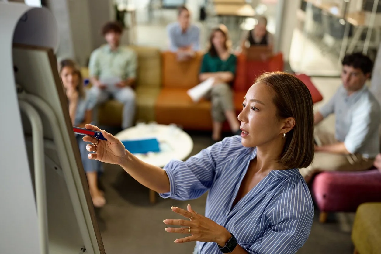A woman giving a presentation to a group of people in a casual office setting, using a whiteboard or flip chart.