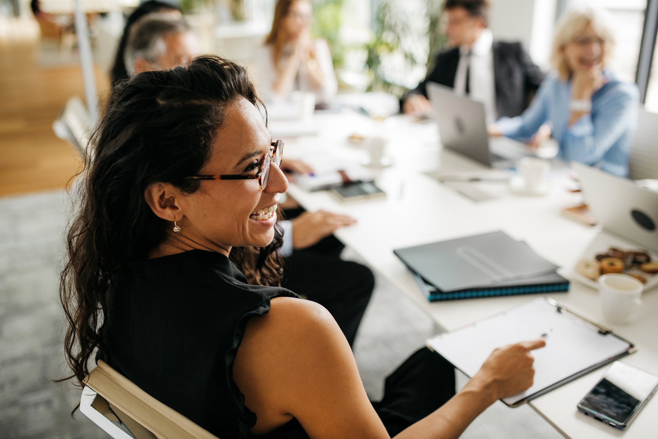 A woman with dark curly hair and glasses smiling during a meeting in a bright office with other blurred people in the background.