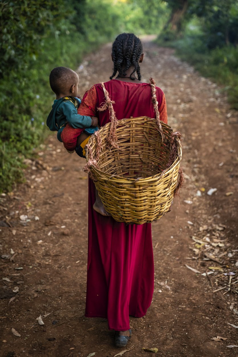 Woman wearing a red dress walking on a dirt road while carrying a large empty wicker basket on her back and holding a small child in her arms. The surrounding area is lush with greenery.