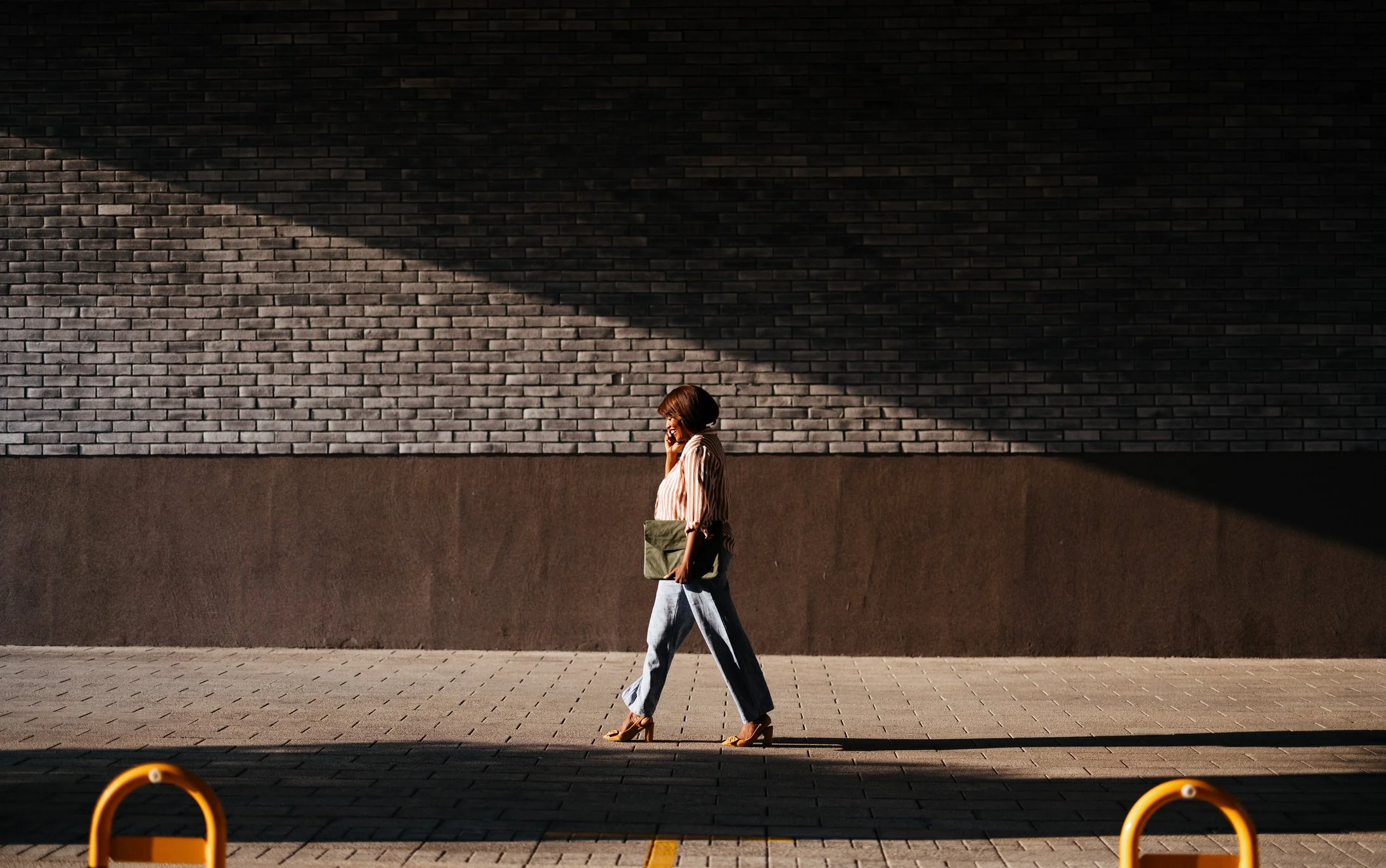 A woman walking along a sidewalk, talking on her phone, carrying a bag, with shadows cast on a brick wall.