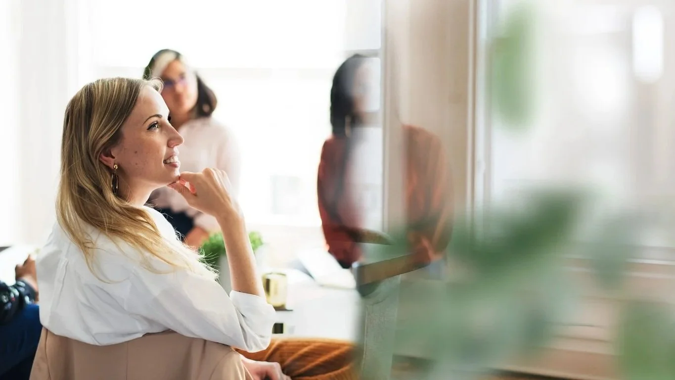 A group of women in a bright office space, with one woman in focus smiling and looking to the left, others blurred in the background near a window.