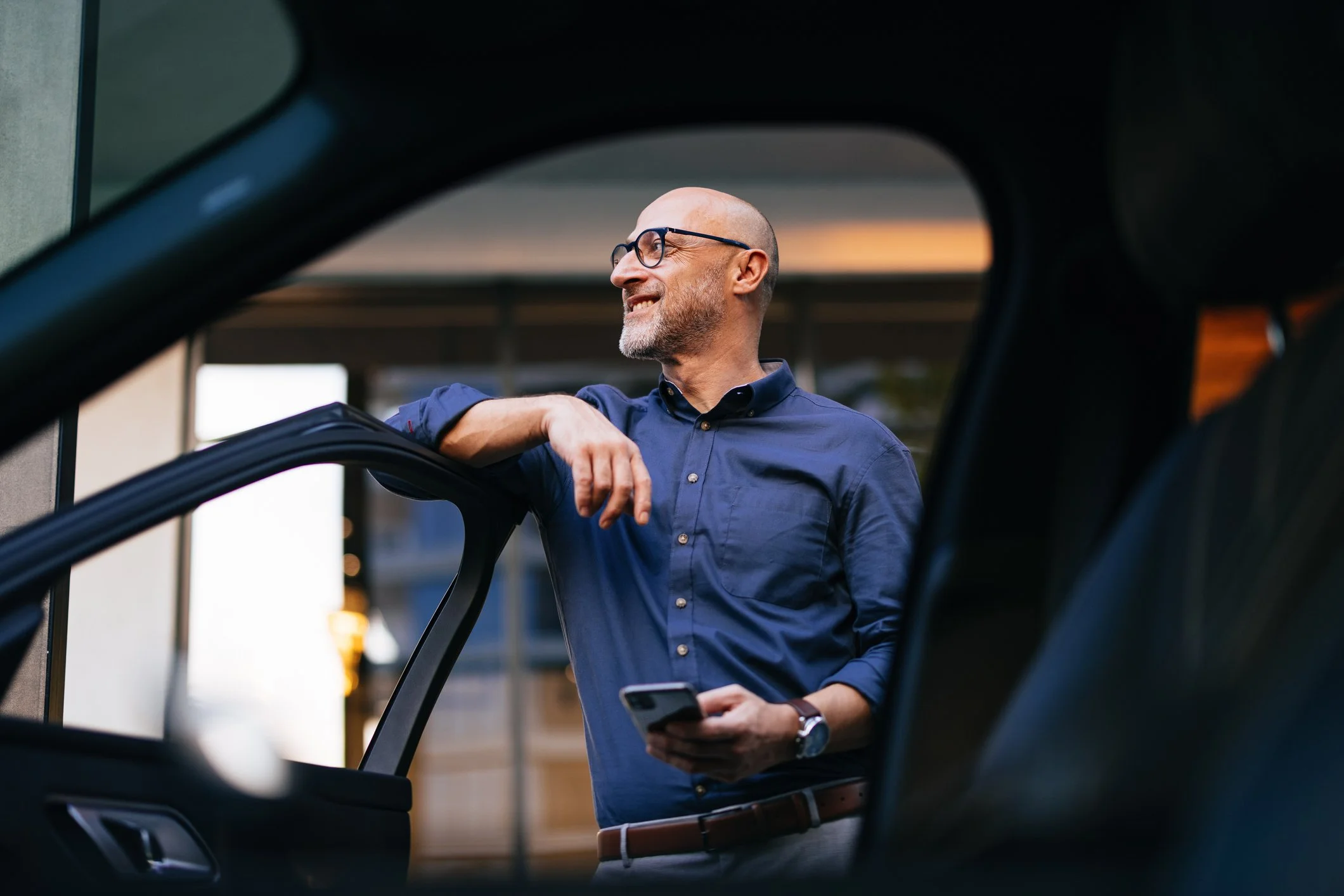 A man with glasses and a beard is standing outside a building, smiling and looking to his right, while holding a smartphone in his left hand. The photo is taken from inside a vehicle, with the vehicle's window or frame partially visible in the foreground.