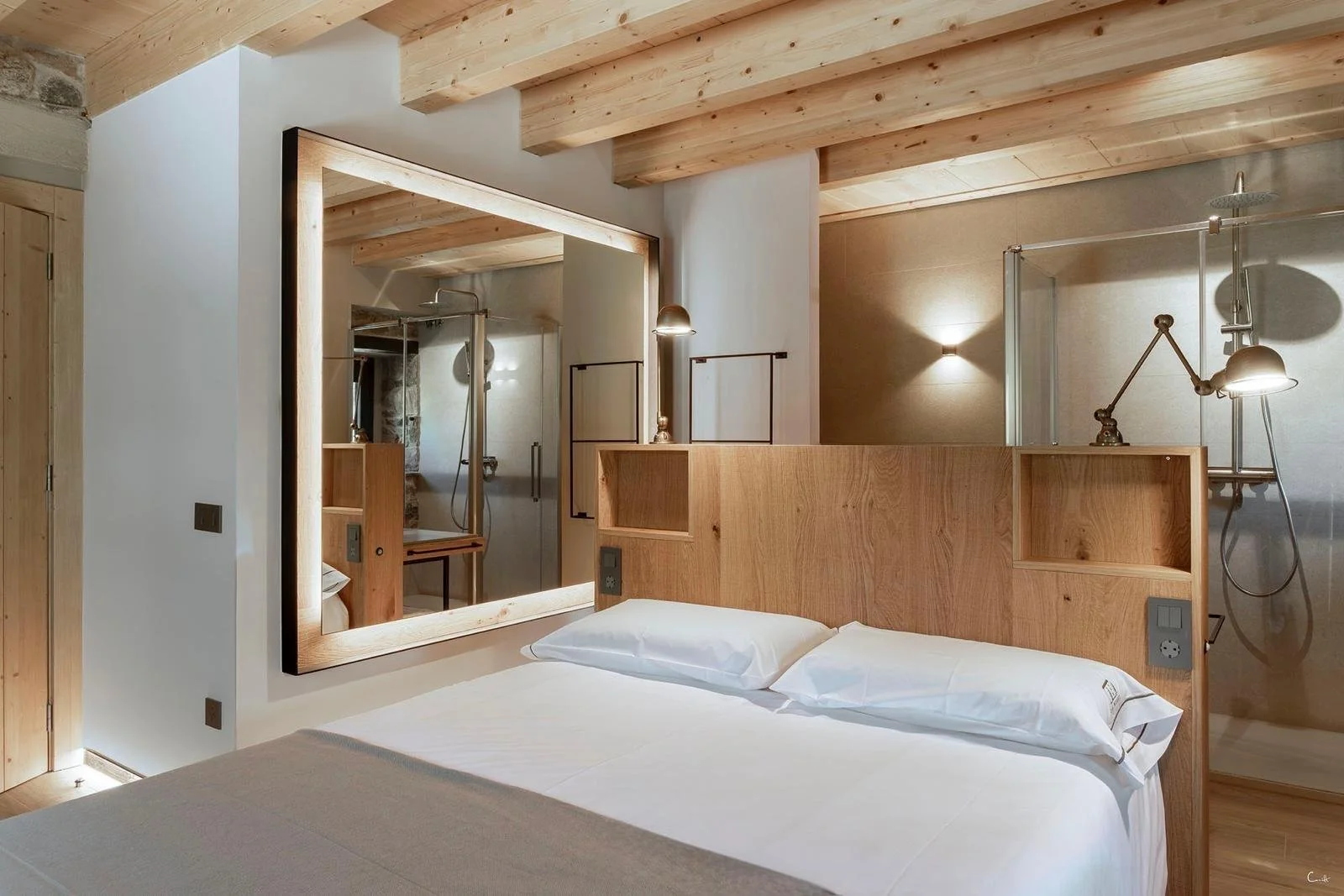 Habitación moderna con cama doble, paredes blancas, techo de madera, y baño con ducha y lavabo visible a través de un espejo grande.