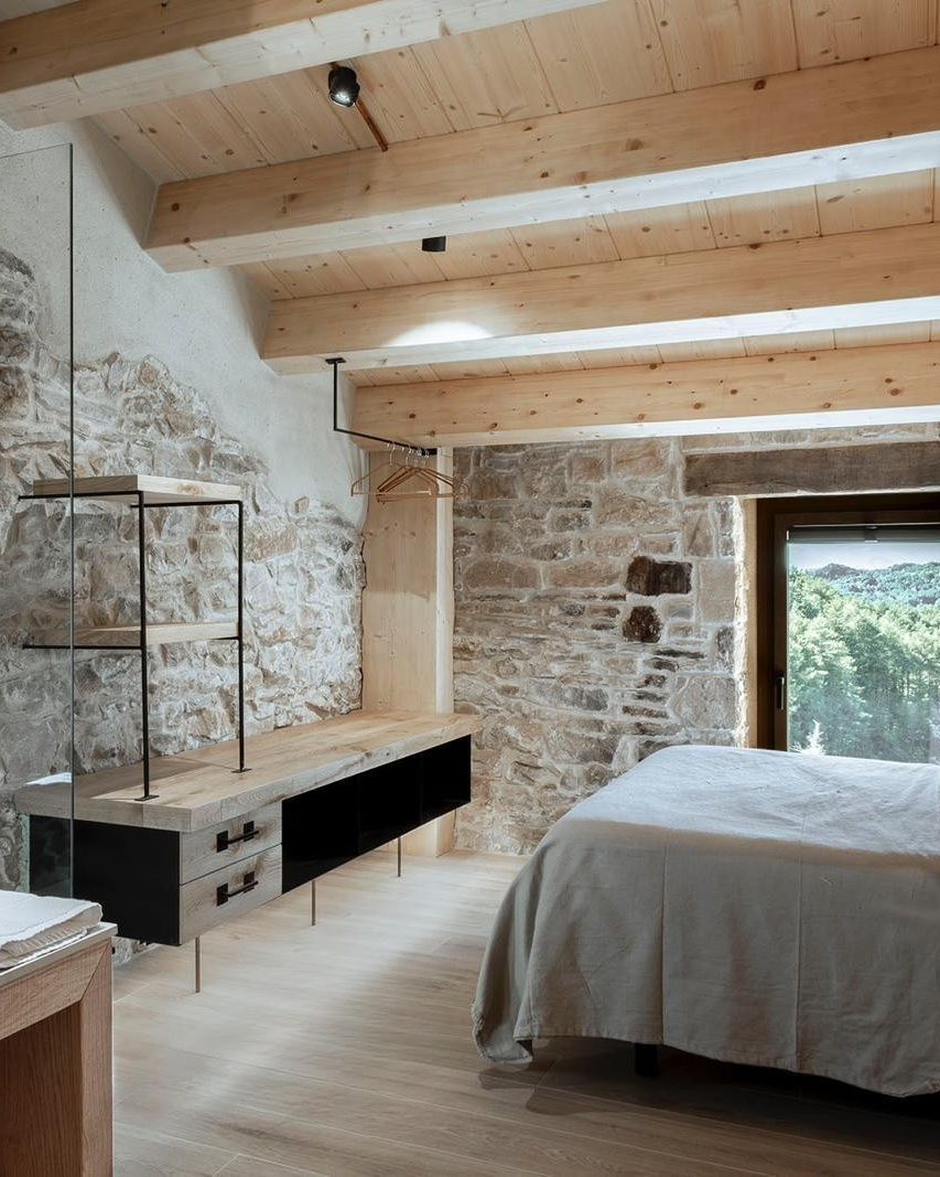 Habitación moderna con paredes de piedra, techo de madera y vista a la naturaleza desde la ventana.