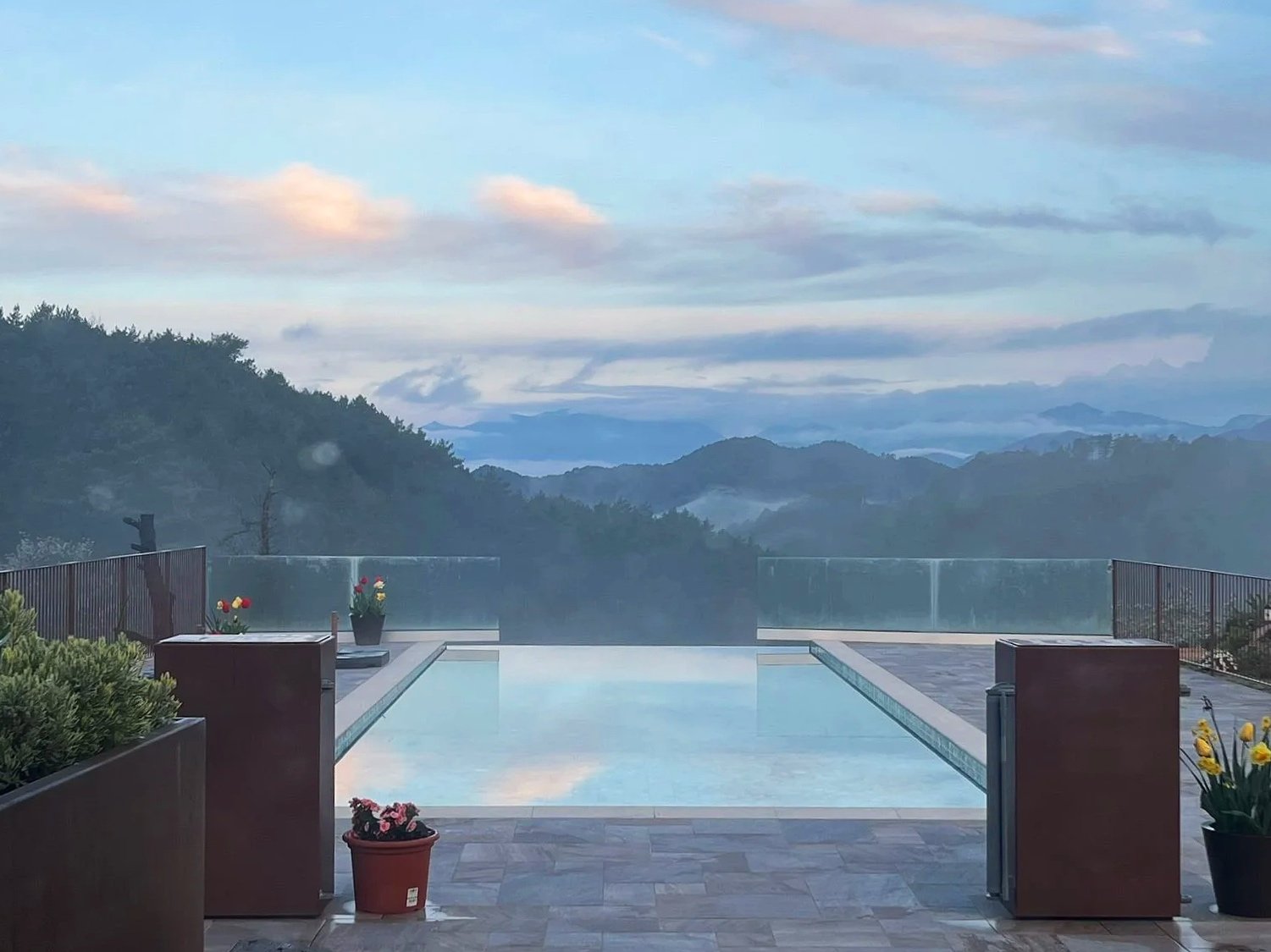 Piscina con vista a las montañas y cielo nublado, rodeada de plantas y flores en macetas de barro y madera en un día húmedo y fresco.