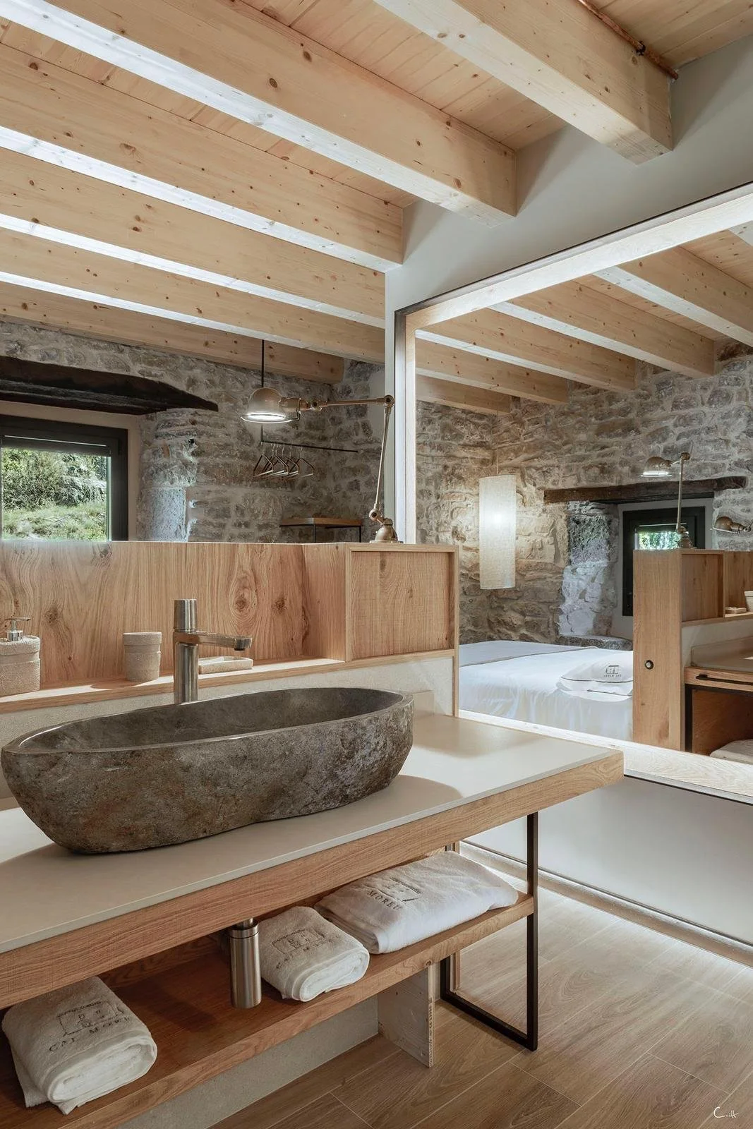 Interior de una habitación con paredes de piedra, techo de vigas de madera, lavabo de piedra en encimera de madera y cama en el fondo.