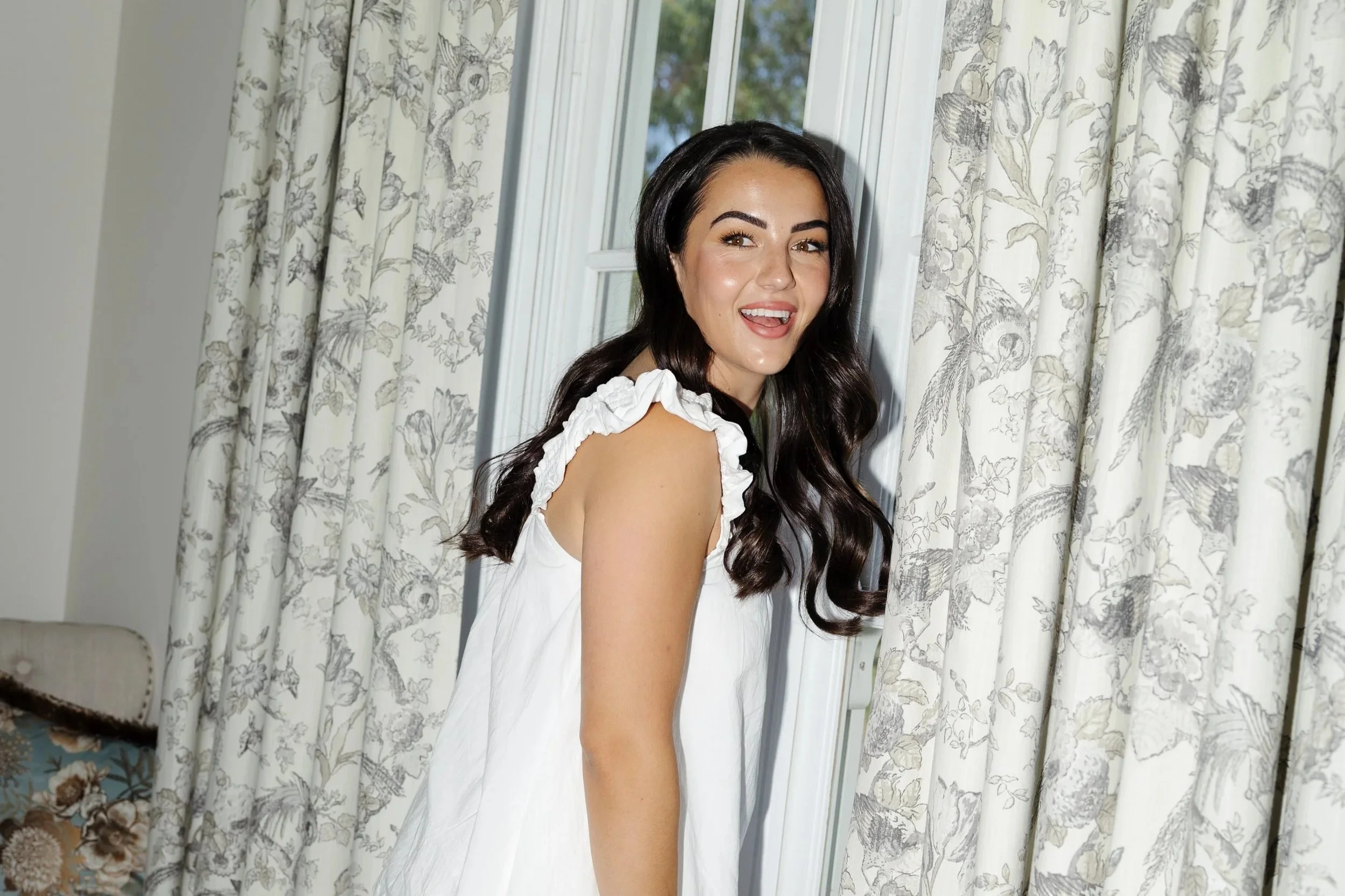 A young woman with long dark hair wearing a white dress with ruffled shoulder straps, standing indoors near a window with floral curtains, smiling and showing her tongue.