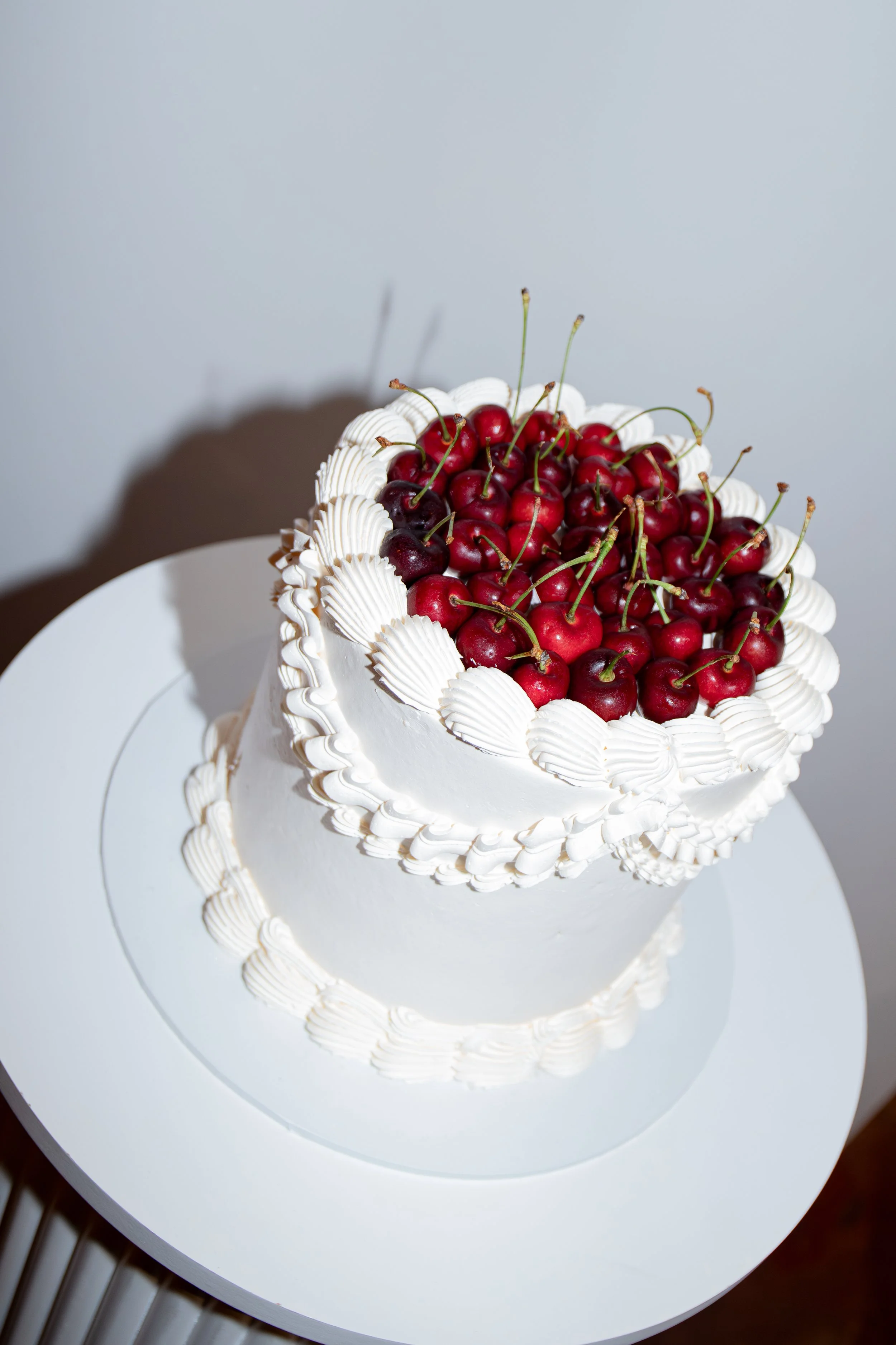 A white birthday cake decorated with cherry toppings and white piping, placed on a white cake stand.